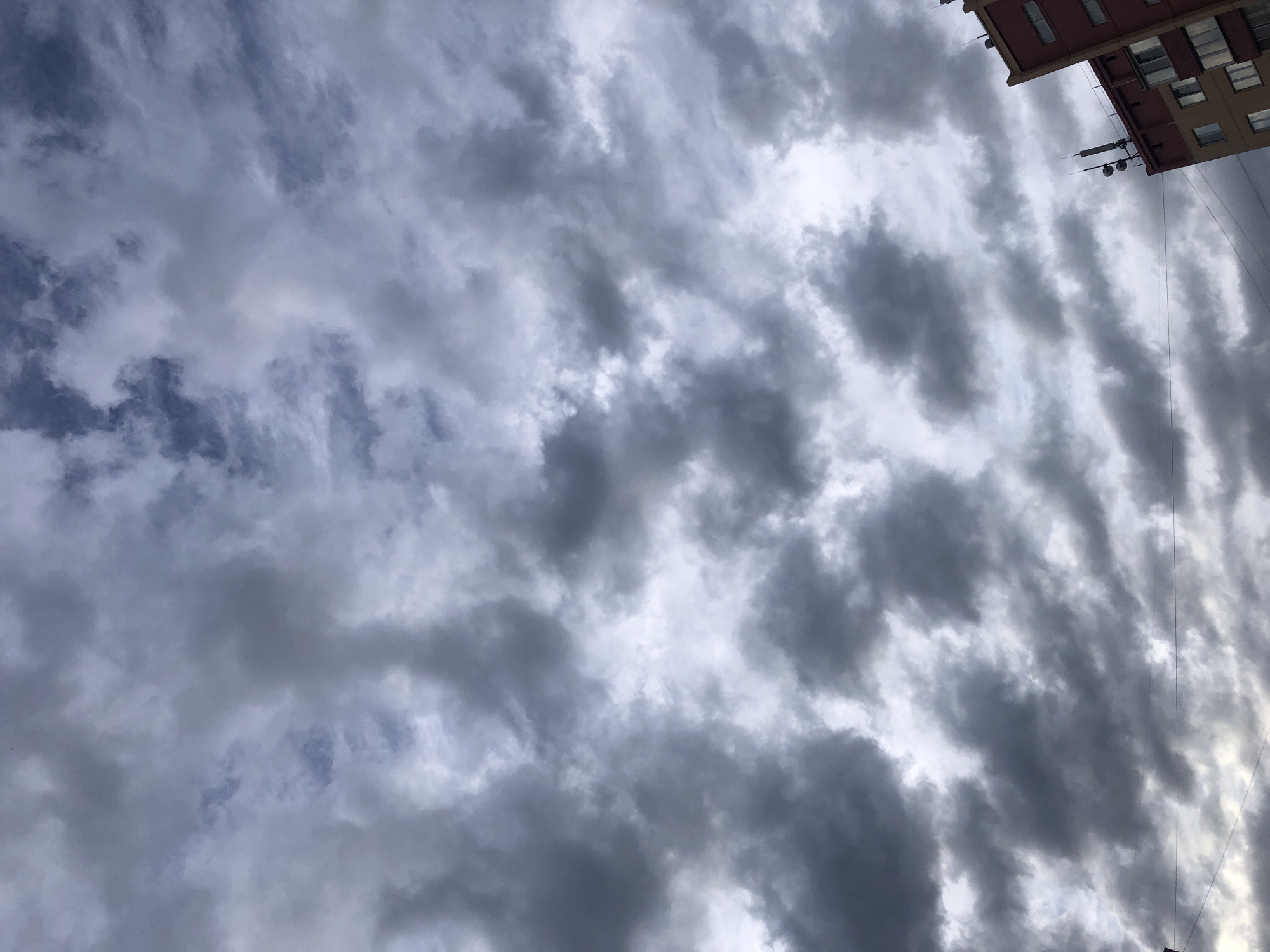 The image depicts a cloudy sky, likely taken in an urban environment. Altocumulus clouds are visible, characterized by their patchy and layered appearance. A portion of a building is seen in the upper right corner, indicating the presence of city structures. Electrical wires are also visible.
