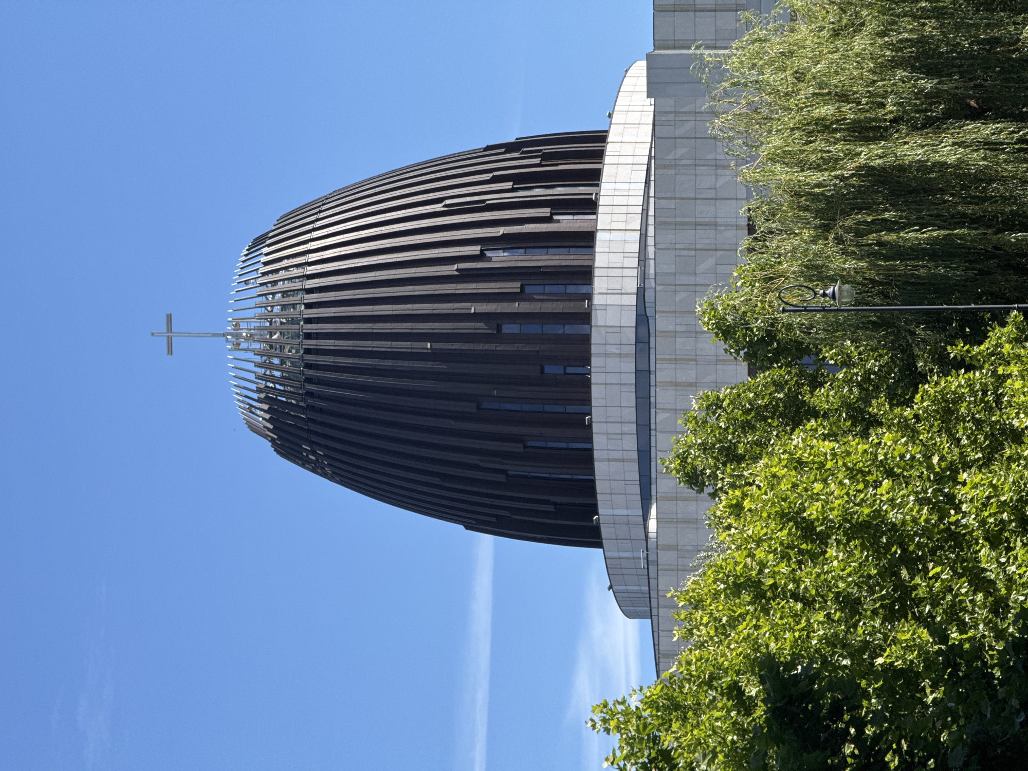 This image depicts a modern architectural structure, likely a church or religious building, in Warsaw, Poland. The prominent feature is a large, oval-shaped building with a dark, slatted exterior. A metallic cross is visible at the very top of this structure. Adjacent to the curved building is a taller, lighter-colored rectangular building with a grid-like facade. Lush green trees, one appearing to be a weeping willow, partially obscure the buildings, suggesting a park-like or landscaped setting. A black lamppost with a decorative light fixture is also visible amongst the foliage.
The scene is set against a clear, bright blue sky, indicating daytime, likely morning or afternoon, with sunny weather. The lighting suggests a fair amount of sunshine, casting shadows and highlighting the textures of the buildings and leaves. The perspective is from a low angle, looking up at the architecture and sky. There is no visible text in the image. The overall impression is one of modern design integrated with nature in an urban environment.