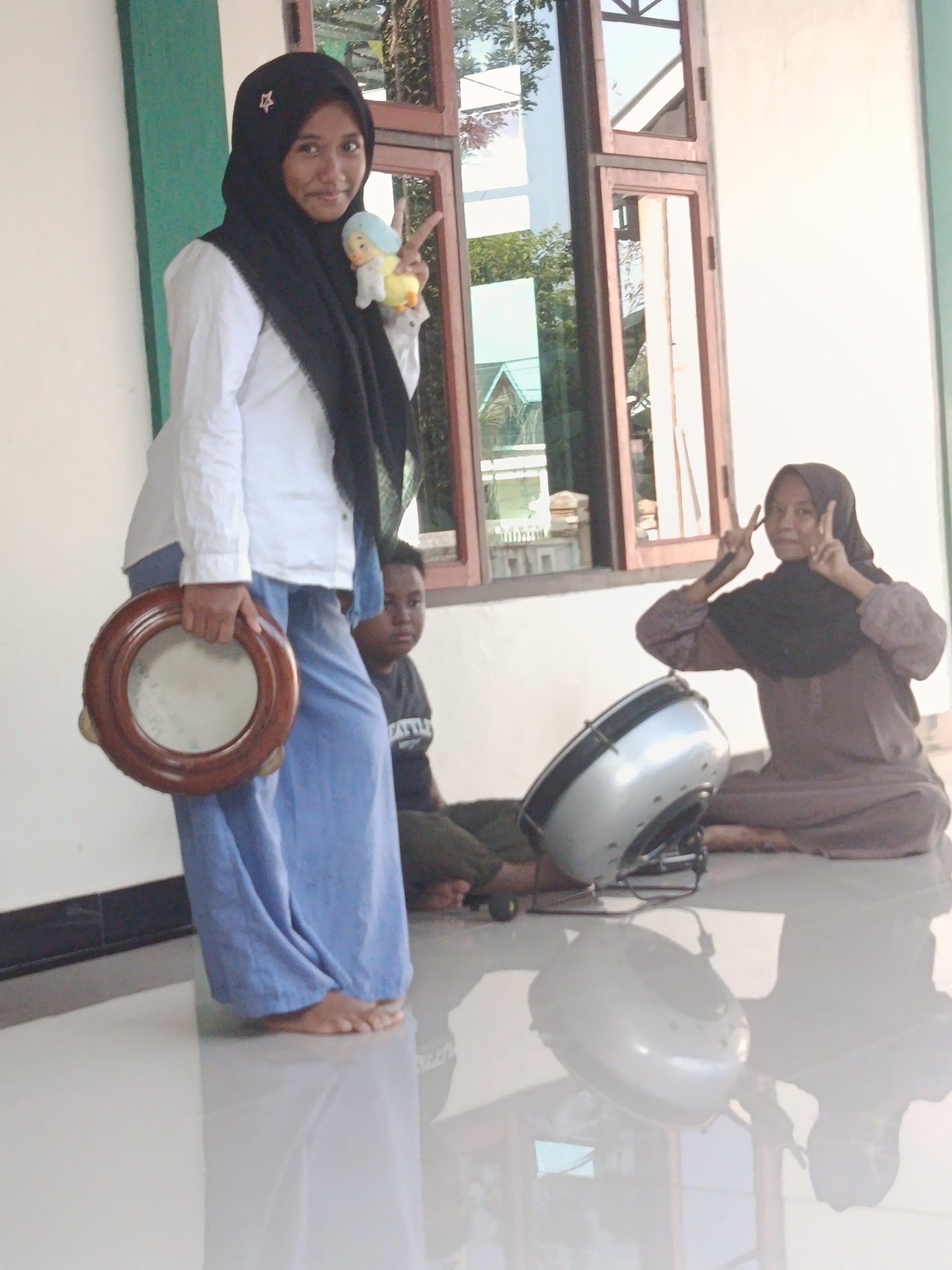 In Swarga Bara, Indonesia, a young woman wearing a black hijab and a white shirt stands on a glossy tiled floor, holding a tambourine with a wooden frame. She is smiling and holding up a small doll with her right hand, making a peace sign with two fingers. Behind her, to her right, a young boy in a dark t-shirt is seated. Further to the right, another young woman in a brown hijab is also seated, making a peace sign with her fingers. A large silver drum is positioned in front of the second seated woman. The background features a building with large windows and a green wall section on the left. The scene appears to be outdoors or in a transitional space between indoors and outdoors, with natural light illuminating the area.