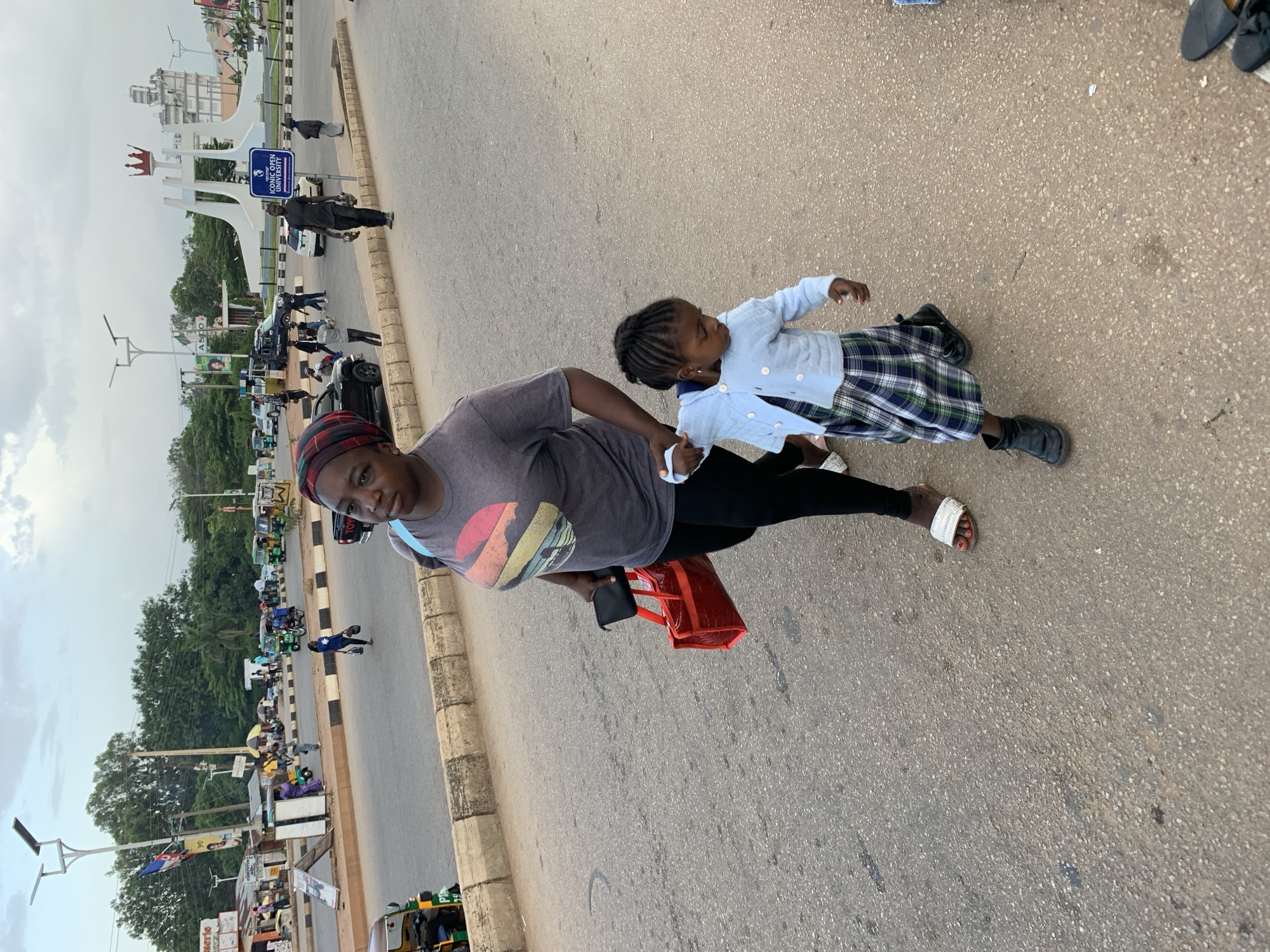 The image shows a street scene in Jos, Nigeria, with a woman and a young girl in the foreground. The woman is wearing a grey t-shirt with a colorful circular design, black leggings, and a headscarf with red and black checkered patterns. She is holding a black clutch and a red shopping bag. The young girl is wearing a light blue cardigan over a plaid skirt and black shoes. She is holding the woman's hand. They are walking on a paved road next to a sidewalk.
In the background, there is a large white sculpture with a crown on top, a sign that reads "ICONIC OPEN UNIVERSITY", and several vehicles, including tricycles (Keke Napep) and cars, lined up along the road. Trees and buildings are visible further in the distance. The sky is overcast, suggesting a cloudy day. The scene appears to be bustling with activity, as indicated by the presence of multiple people and vehicles. The overall impression is of a typical day in an urban environment in Nigeria.