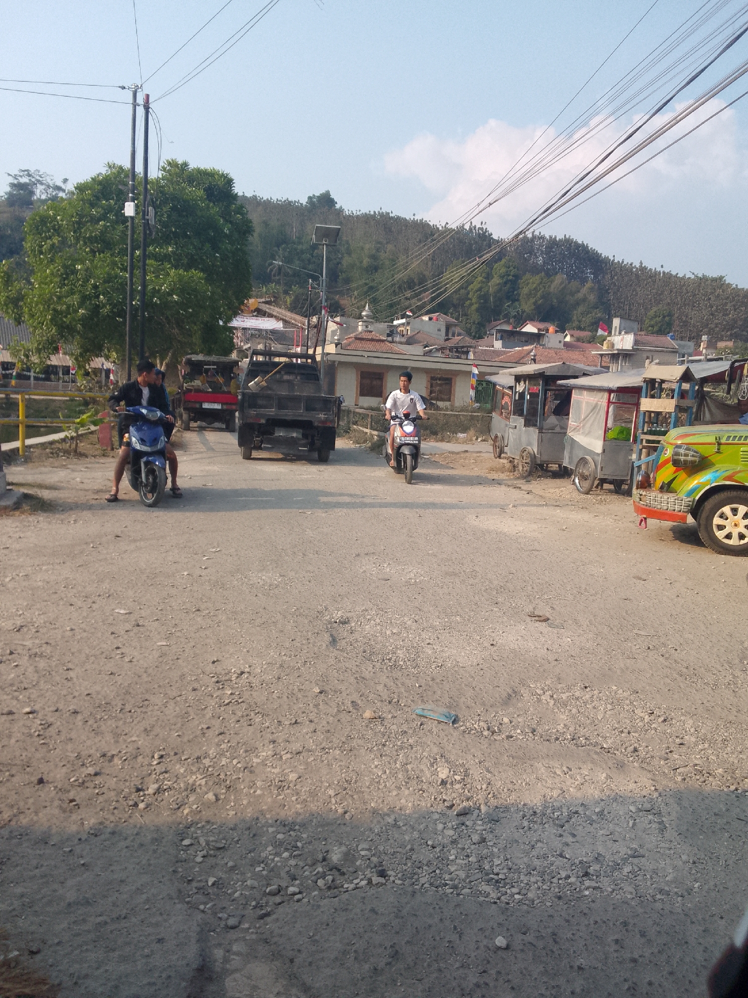 The image depicts a dirt road in what appears to be a rural or semi-urban area, likely Ciburuy, Indonesia, given the context. The road is unpaved and uneven, with scattered stones and debris. In the foreground, a dark shadow is cast across the road.
Several vehicles are present. To the left, a person is riding a blue motorcycle, with another person sitting behind them. Further down the road, a black truck is parked, and behind it, a red truck is visible. To the right, there are several stationary carts, possibly food vendors or small shops, with roofs. One of these carts is partially obscured by a brightly colored, small, open-sided vehicle that resembles a modified jeep or buggy. Another person is riding a scooter on the right side of the road, heading away from the camera.
The background shows a dense, treed hillside dotted with buildings, suggesting a populated area nestled into the landscape. Power lines crisscross the sky above the scene. The sky is mostly clear with some scattered clouds, and the lighting indicates it is daytime, likely late afternoon due to the long shadows. The overall impression is one of everyday activity on a local road.