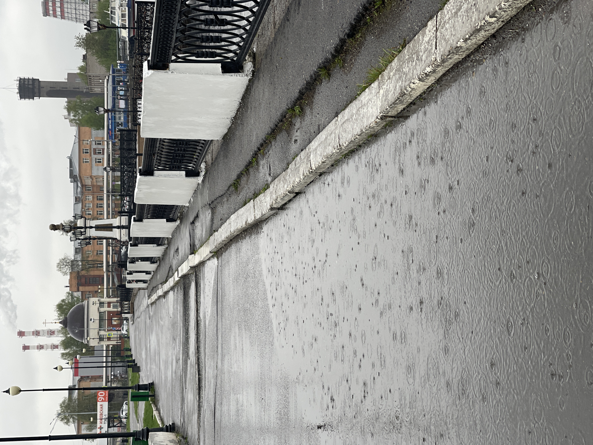 The image depicts a street scene in Izhevsk, Russia, on a rainy day. The foreground is a wet concrete embankment with rain droplets creating ripples in the standing water. A weathered concrete curb runs along the edge of the embankment, with patches of green vegetation growing between the curb and the adjacent asphalt surface.
A black wrought-iron fence with white concrete supports lines the side of the asphalt surface away from the embankment. Beyond the fence, a row of streetlights with black metal poles is visible. Further back, there are buildings of various heights and colors, including a building with a dark dome and several industrial chimneys. A water tower is also visible in the background.
A sign with the number 90 is visible. The scene is overcast and appears to be taken during the daytime.