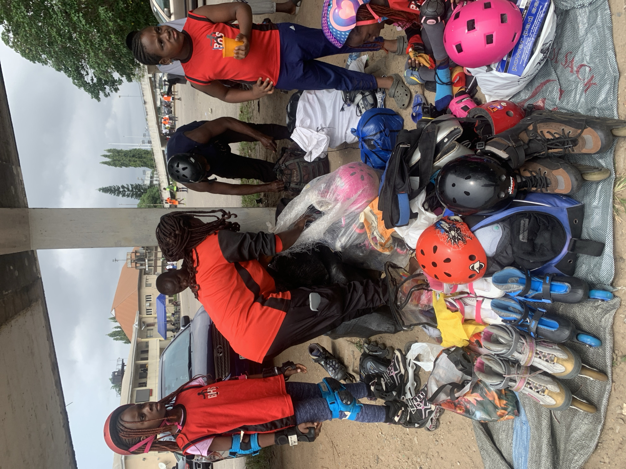 In Alagbede, Nigeria, under a concrete overpass, a group of people are preparing for rollerblading. Two young girls, dressed in protective gear and vests, stand amongst a collection of rollerblades, helmets, and bags. One girl wears a red vest with "BHS" printed on it, while the other sports a red vest with a heart design helmet. An adult, wearing a dark helmet and a black shirt, is bent over a bag, seemingly organizing equipment. The ground is sandy and littered with various items, including a car parked behind the group. The scene is likely a casual gathering or practice session for rollerblading.