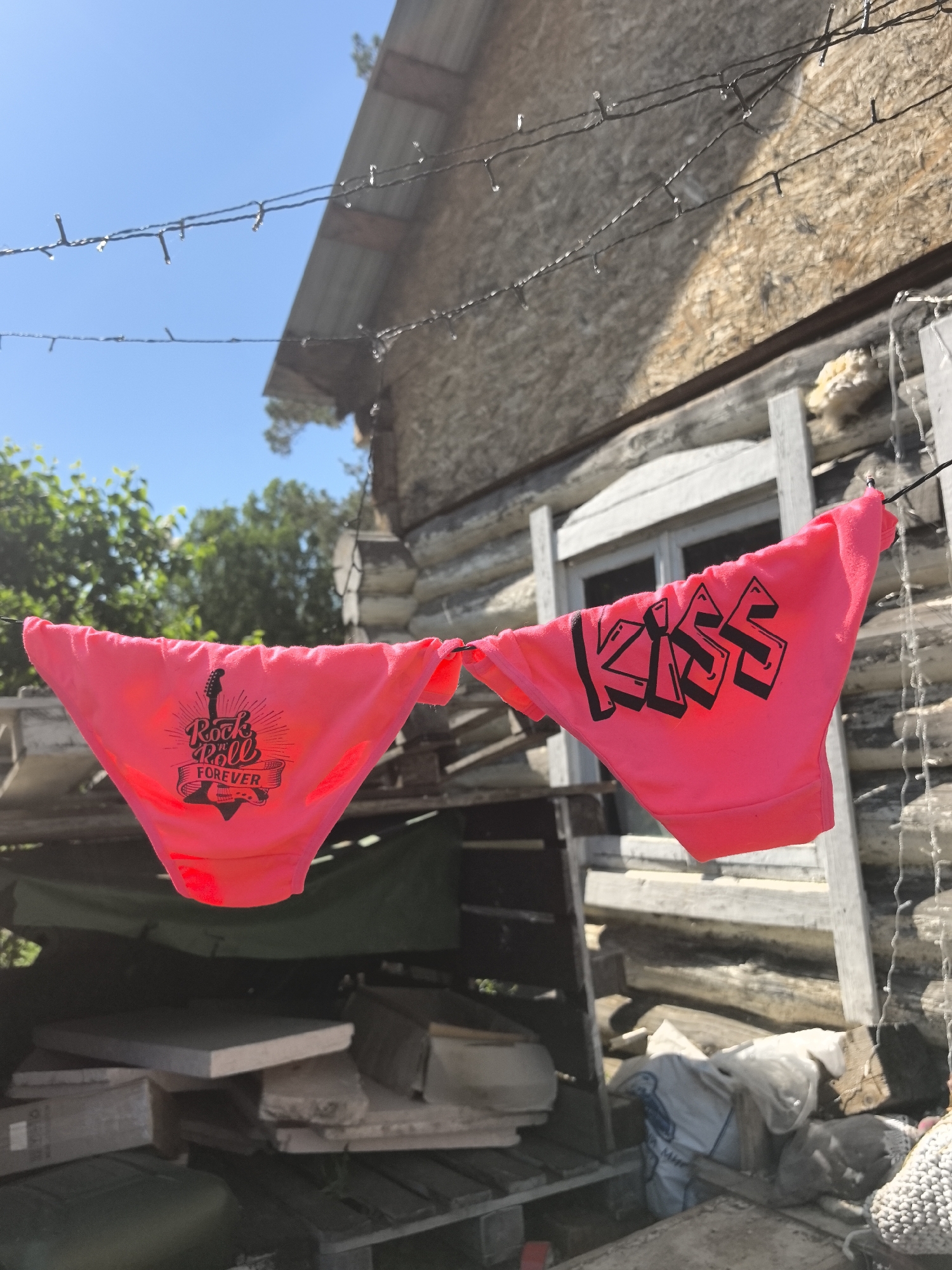 A pair of bright pink panties hang on a string against the backdrop of a rustic, wooden building. The left panty features a graphic of a guitar and the words "Rock & Roll Forever." The right panty displays the word "KISS" in a stylized, black font. The background includes a partly-visible window, a wooden pallet with assorted objects, foliage, and a clear blue sky. The building appears to be located outside, with strings of small lights hanging nearby. The scene may be in Noviy, Russia.