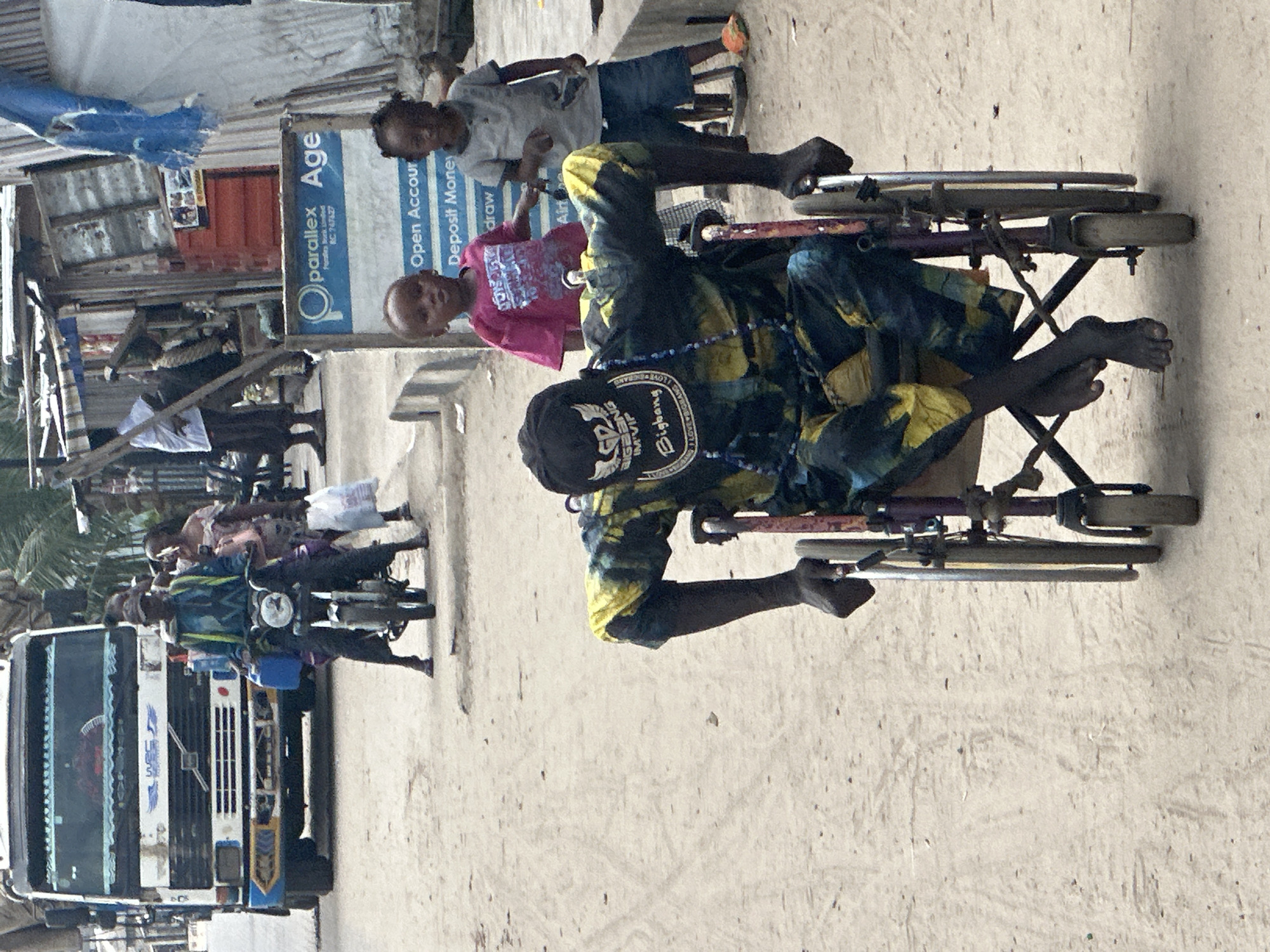 In a dusty street scene in Badagry, Nigeria, a man in a wheelchair is the central focus. He is dressed in a vibrant yellow and black patterned outfit and wears a black baseball cap with a circular logo. The wheelchair appears to be well-used, with visible wear and tear.
In the background, several other individuals are present. Two young children, a girl in a pink t-shirt and another in a grey t-shirt and blue shorts, stand near a blue and white sign that reads "Parallex Age" with text about opening accounts and depositing money. Further back, a person on a motorcycle, wearing a blue and green jacket, is stopped by the side of the road. A large white truck is also visible on the left side of the frame. Buildings with corrugated metal roofs and canvas awnings line the street. The ground is uneven and sandy, indicative of a less developed urban area. The overall impression is a snapshot of daily life on a busy street.