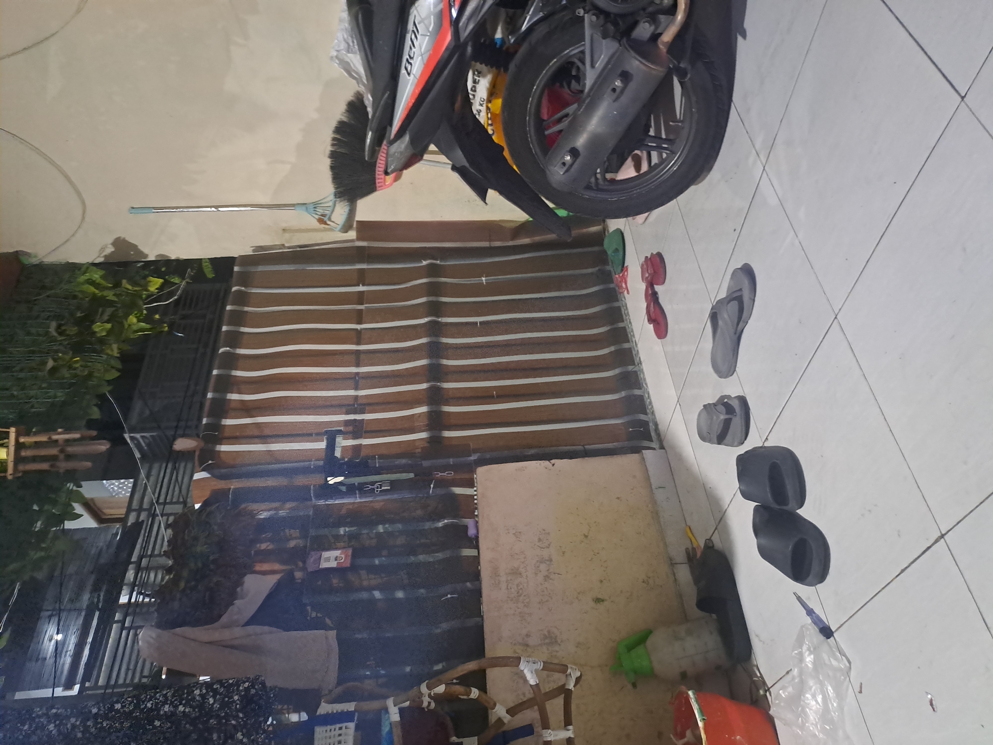 The image depicts an outdoor scene at night in Kebonagung, Indonesia, likely a residential area. In the foreground, the tiled floor is scattered with various footwear, including a pair of black slides, a pair of grey flip-flops, and a pair of red flip-flops. A red bucket and a green spray bottle are visible on the left side of the image. To the right, the rear section of a black and red motorcycle is parked. Behind the motorcycle, a broom with a blue handle rests against a light-colored wall. A striped brown and beige awning or curtain hangs on a structure in the middle ground. The background features a dark, cluttered area with some greenery and structural elements, suggesting a yard or alleyway. The lighting suggests it is nighttime.