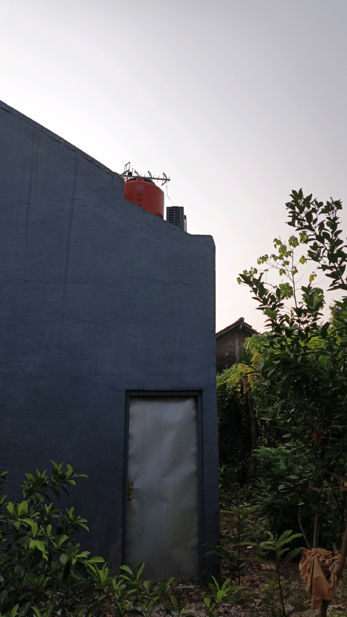 The image depicts an exterior view of a gray building with a metal door. An orange water tank is visible on the roof of the building, along with an air conditioning unit. Lush green foliage surrounds the building, suggesting a somewhat overgrown or natural setting. In the background, glimpses of a tiled roof and more dense vegetation can be seen. The lighting indicates it might be late afternoon or early morning, given the warm tones of the sky. The scene appears to be located in a residential or semi-urban area of Cikal, Indonesia, characterized by its buildings and abundant plant life. There are no people or animals present in the image.