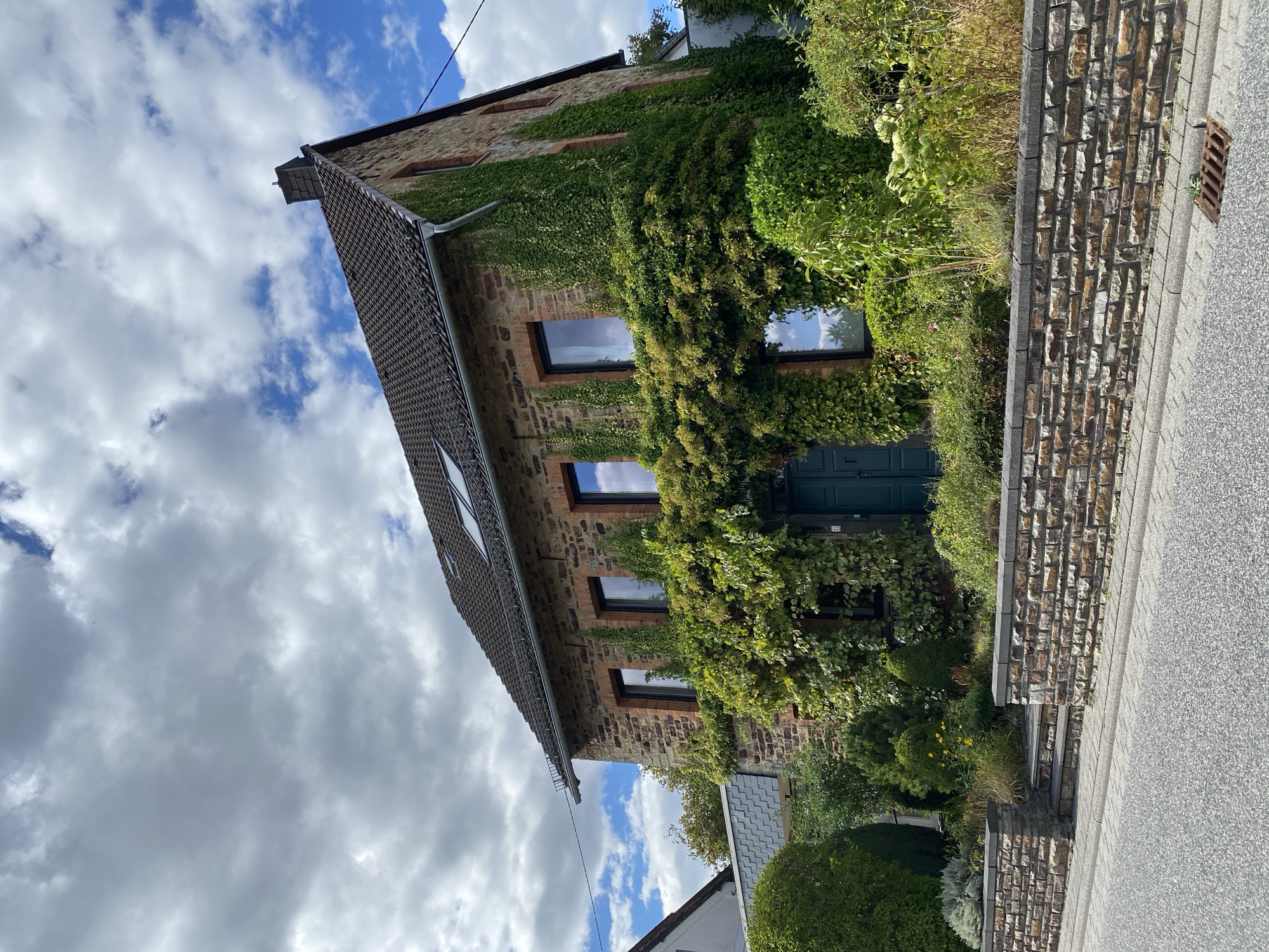 The image shows a two-story house in Flammersfeld, Germany, with a stone facade partially covered in ivy. The house has a dark tiled roof with a chimney and several windows with dark frames. A dark blue door is visible at the front entrance, which is surrounded by dense green foliage. The property is bordered by a stone wall and a paved sidewalk, with a street in the foreground. The sky is partly cloudy with blue patches. The scene appears to be a residential area, with no people or vehicles visible.