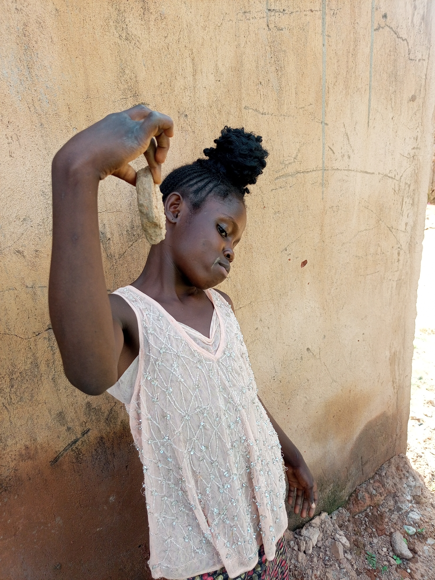 A young Black girl, with braided hair styled in a bun, stands against a tan, textured wall, holding a light-colored rock in her right hand, raised above her head. She wears a light pink sleeveless top adorned with a diamond-patterned overlay and sequins. Her gaze is directed downwards and to her right. The wall, which appears to be a building exterior, shows some cracks and dark marks. The ground in the foreground is uneven, composed of dirt and small stones, with a few green sprouts visible. The lighting suggests it is daytime, with sunlight casting subtle shadows. The scene is set in an outdoor environment, likely in the city of Tempure, Nigeria.