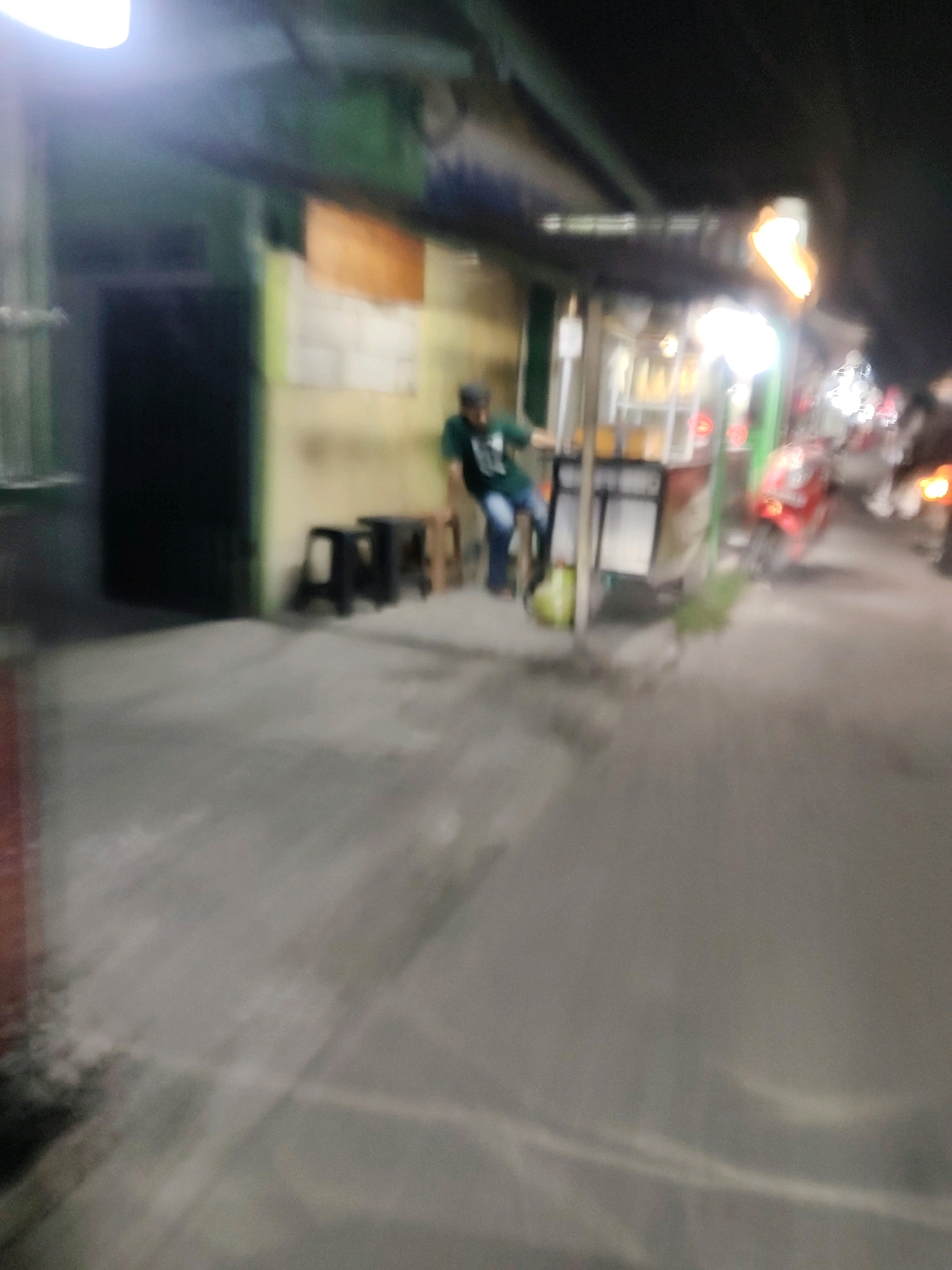 The image depicts a street scene at night in Mekarmukti, Indonesia. In the foreground, a paved road is visible, reflecting the ambient light. To the left, a building with a dark entrance and a green facade stands adjacent to a wall with orange and yellow paneling. In front of the building, several dark stools are arranged. A person wearing a green shirt and blue pants is seated on one of the stools, leaning forward. Behind the person, a street food stall is illuminated, featuring a brightly lit display with yellow contents. Various vehicles, including a red motorcycle, are present in the background, suggesting a moderately busy street. The overall scene is blurry, likely due to low light conditions and movement during the capture.