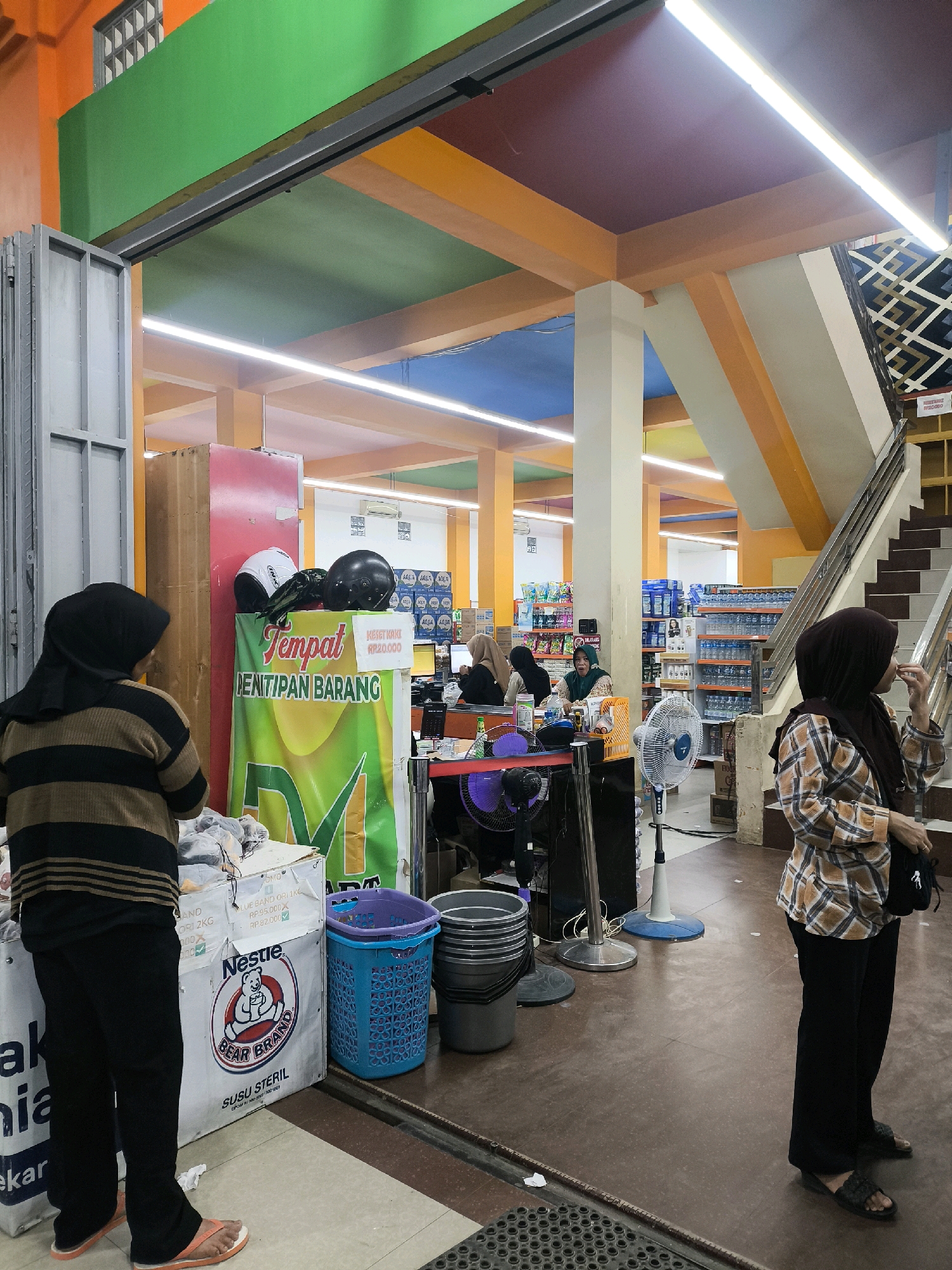 The image depicts the interior of a brightly colored supermarket or general store, likely located in Posso, Indonesia, given the context of the provided crops. The ceiling features a vibrant, segmented design in orange, green, and blue. A central support column is painted white.
In the foreground, a person wearing a black hijab and a horizontally striped long-sleeved shirt and black pants stands with their back to the camera, facing an area where items are displayed. To their left, a large white box with the "Nestle Bear Brand Susu Steril" logo is visible, along with a blue plastic basket and stacked gray buckets.
Further into the store, several people are seen. Two individuals are positioned behind a counter. One is wearing a green hijab, and another a light-colored hijab. Another person in a dark hijab is visible near a staircase to the right. The person on the far right of the image wears a plaid long-sleeved shirt, black pants, and a dark hijab. They are looking to the left and holding something to their mouth. A white oscillating fan stands to the left of this person.
Shelving units stocked with various products, including bottled water, are visible in the background. A sign with the text "Tempat Penitipan Barang" (Luggage Deposit Area) is prominently displayed on a green banner with yellow accents, with two helmets placed on top of a red and brown structure behind it. Another sign indicates "KESEI KAKI RP20,000," suggesting a charge for foot care items or services. The floor is tiled with a brown and beige pattern. The overall impression is that of a busy retail environment.