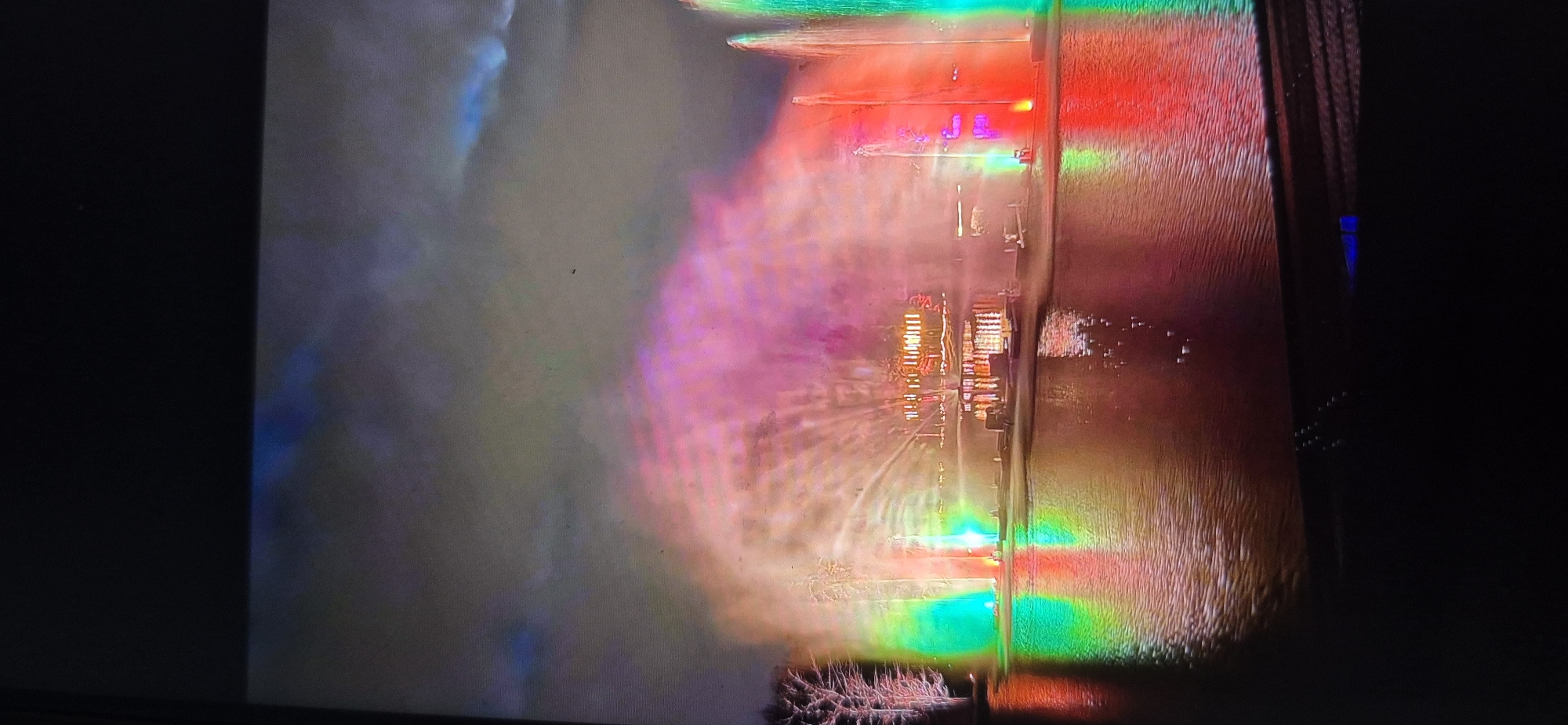 An aerial or high-angle photograph captures a brightly illuminated structure, likely a bridge or pier, extending horizontally over a body of water at night. The scene is heavily enveloped in haze, fog, or smoke, causing the intense artificial lights to diffuse into broad, vibrant swaths of color.
The central structure is adorned with various colored lights, predominantly red and green, creating strong reflections on the water below. Prominently displayed on the structure, neon-like pink or purple lights clearly spell out "JL" or "J.L.". Nearby, clusters of warm yellow and orange lights suggest buildings, containers, or other features. The thick haze on the left side of the image glows with diffused blue and purple light, transitioning into pink and red hues closer to the central structure.
On the far right, a dark vertical element, possibly a structural support or part of a camera's frame, features a small, distinct blue light. In the bottom-left corner, an intricate, dark, and possibly natural pattern, resembling vegetation, is partially visible through the colorful haze. The water's surface exhibits texture and ripples, enhancing the reflections. No people are visible. The time of day is night, and the notable atmospheric haze creates a dramatic and ethereal ambiance.
