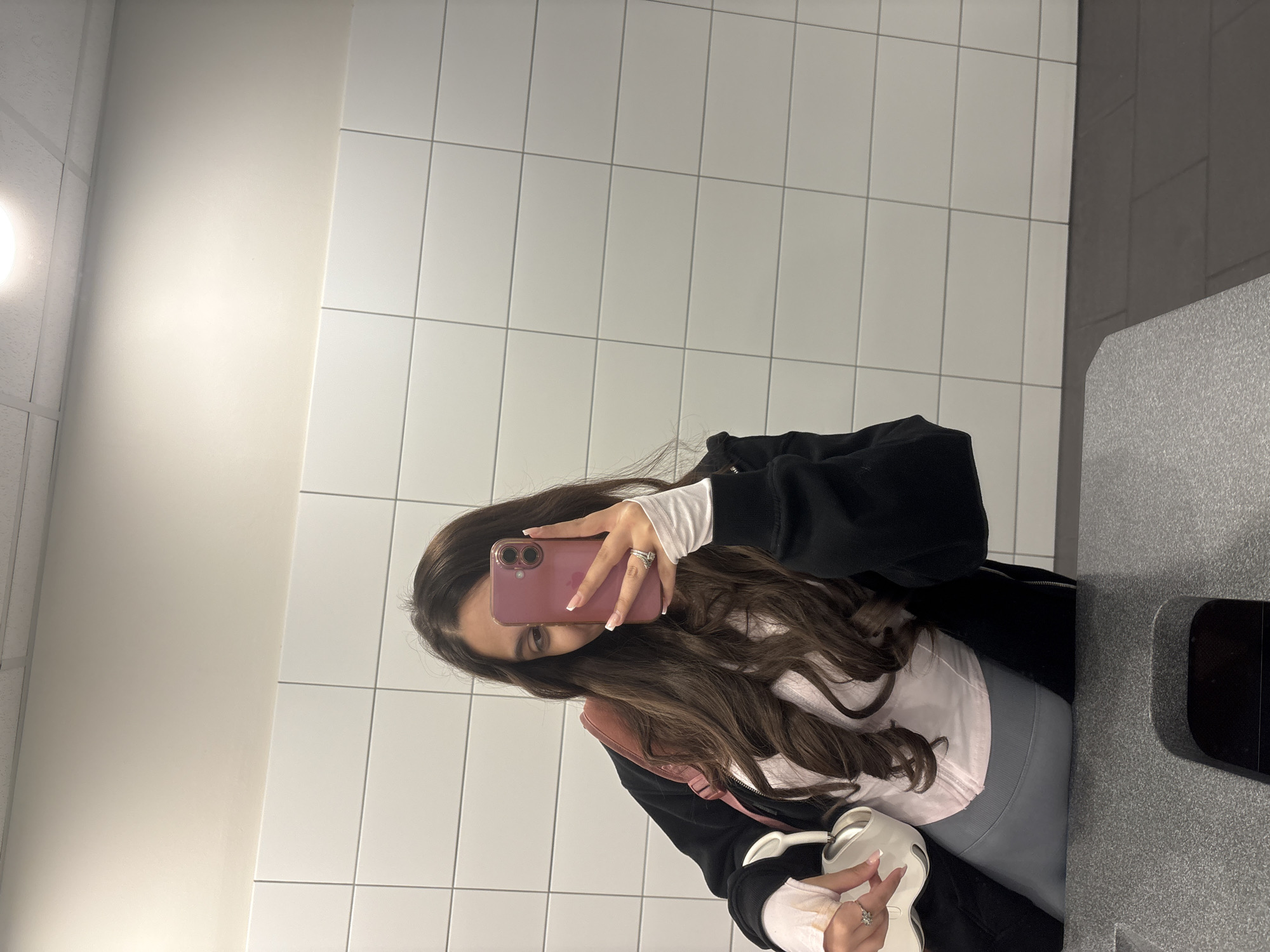 A person is taking a selfie in a bathroom. The person is holding a pink smartphone in their right hand, partially obscuring their face. They have long brown hair and are wearing a black jacket over a pink and white layered outfit. The background consists of white tiled walls and a grey countertop. The location is in Mönchengladbach, Germany is unknown based on this image.
