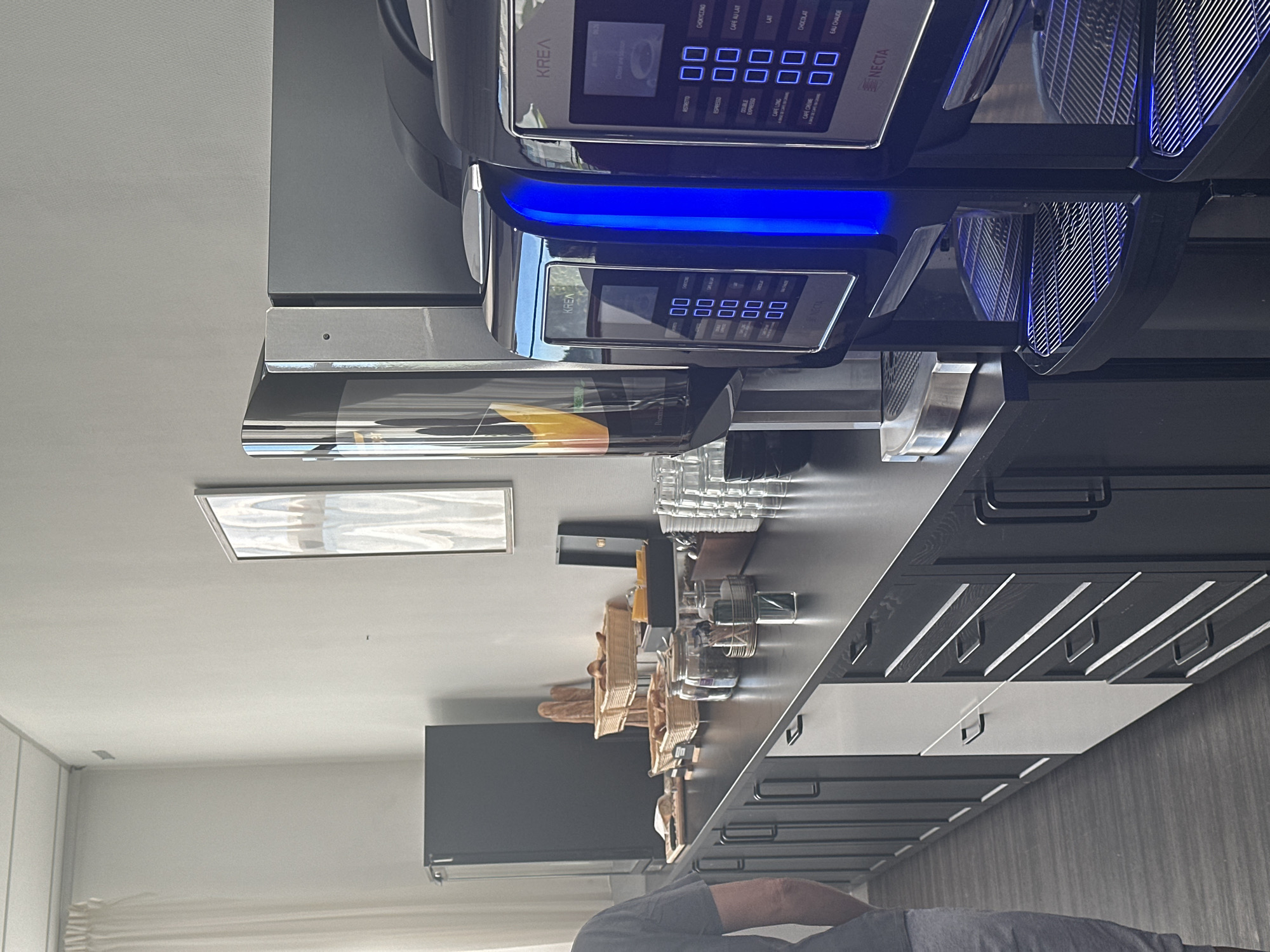 Here is a description of the image:
**Scene:** The image appears to be an interior shot of a kitchen.
**Objects:** Dominating the view is a black and silver coffee machine with blue lighting, branded with "KREA Necta". There are several glass cups and jars on the counter. Baskets contain baguettes and possibly other food items. A television is attached to the wall. A framed picture is on the wall. The kitchen cabinets are black and white.
**People:** Partially visible in the foreground, there is a person wearing a grey shirt.
**Environment:** The kitchen appears modern. There is a window.
**Location:** Assuming all OCR is correct, the location is likely somewhere in Ramonville-Saint-Agne, France.