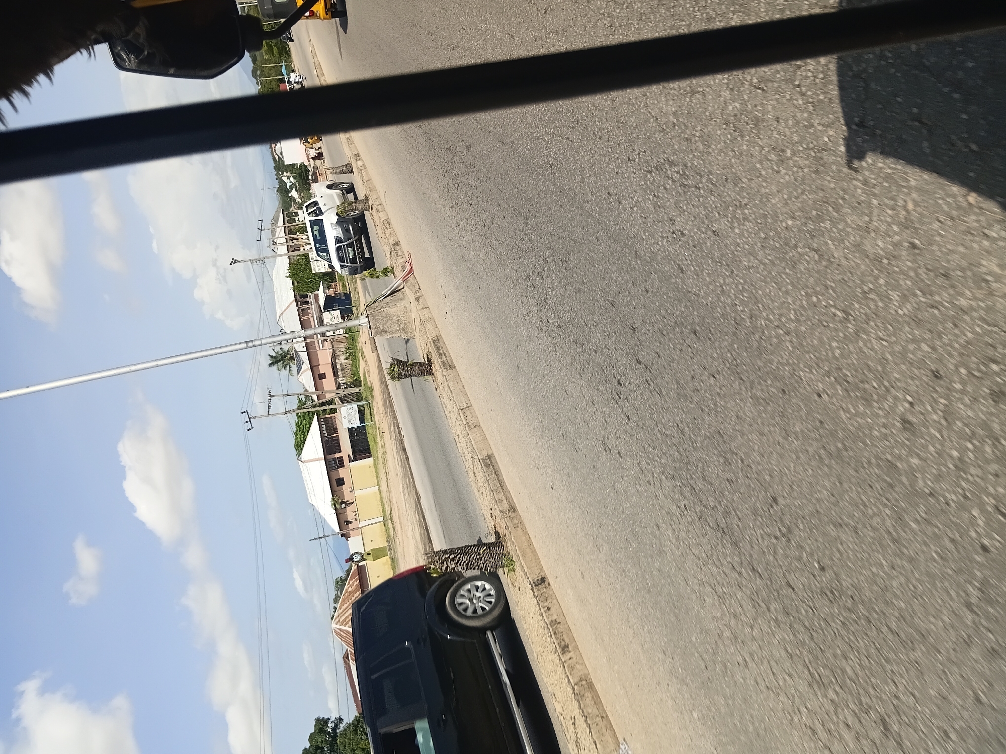 The image depicts a roadside scene in Jalingo, Nigeria, with a focus on the road and surrounding environment. In the foreground, the asphalt road surface is visible, with a portion of a gray concrete curb and planter. On the road, a black SUV is in motion, its rear wheel clearly visible. Further down the road, a white pickup truck is also seen. In the background, several buildings with light-colored walls and white or brown roofs line the street. Electrical poles and overhead wires are present, indicating urban infrastructure. The sky is bright blue with scattered white clouds. The perspective suggests the photo was taken from a moving vehicle, possibly a tricycle or car, as indicated by a dark shadow and a portion of what appears to be a mirror on the right side of the frame. The overall impression is that of a typical daytime scene on a paved road in a Nigerian city.
