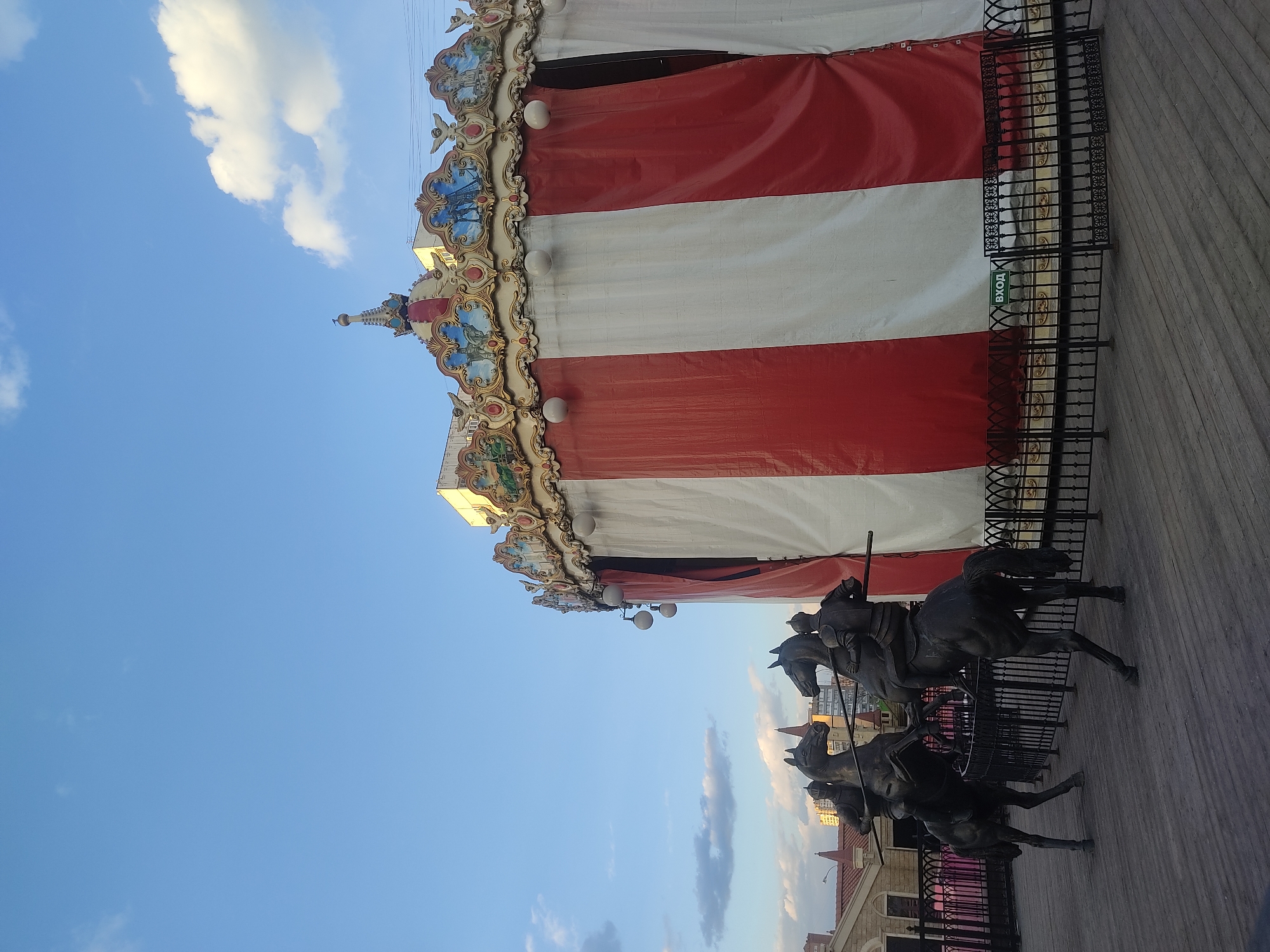 Here is a detailed description of the image:
The image depicts a carousel structure with a red and white striped canopy, ornate decorations, and a sculpture of horse riders in front of it. The location is Moscow, Russia, as evidenced by the inscription "Вход" (meaning "Entrance" in Russian) on a sign near the carousel entrance. The carousel has a decorative border with painted figures and white spherical lights. Below the canopy is a black metal fence with a pattern. In front of the carousel is a sculpture featuring multiple horse riders with spears. The background includes a blue sky with scattered clouds. The image appears to be taken from a low angle.