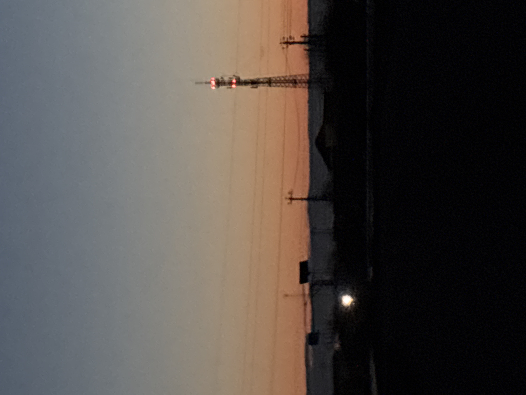 The image depicts a nighttime view in what appears to be a rural or suburban area. There is a communication tower visible in the distance, illuminated with red lights. In the foreground, utility poles and wires are silhouetted against the fading light of the sky. The sky transitions from a light orange near the horizon to a darker blue higher up. The right side of the image is dominated by a dark, almost entirely black, area, likely representing either dense foliage or shadow. Based on the prompt's mention of the city of Прикамский, Russia, it can be inferred the image was taken in or near that location. There is no visual evidence of people, interactions, or specific actions within the scope of the photograph.