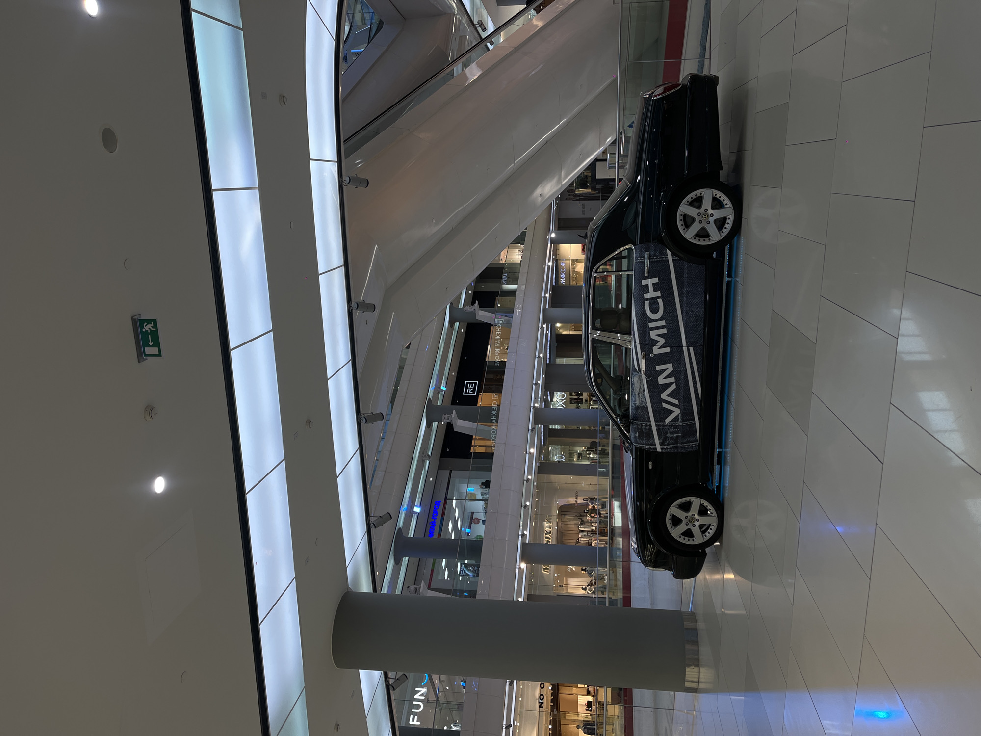 This image captures the interior of a brightly lit, modern multi-level shopping mall in Krasnodar, Russia. The central subject is a dark-colored sedan, prominently displayed on the ground floor, featuring a large "VAN MICH" graphic on its side, resembling a denim texture. The car is equipped with distinctive white multi-spoke rims.
The mall's architecture is sleek and contemporary, characterized by vast white surfaces, luminous panels that run along curved structures (likely housing escalators), and multiple glass-fronted store levels. A wide escalator ascends through the center-left of the frame. Large grey cylindrical pillars punctuate the space. The floor is covered with light-colored rectangular tiles.
Various store names and signs are visible on different levels through the glass railings. Recognizable text includes "FUN," "RE," "SIXTY," "OX," and "MARCA." Other partially visible or mirrored texts from store signs include "СЕЖНУ КОБО" and "CHEMIN KOVE," along with fragments like "НОРОД" and "NO O." A green emergency exit sign with a running figure is affixed to a white wall on the left. The scene suggests a static car display, likely for advertisement or promotion within the bustling commercial environment of the mall.