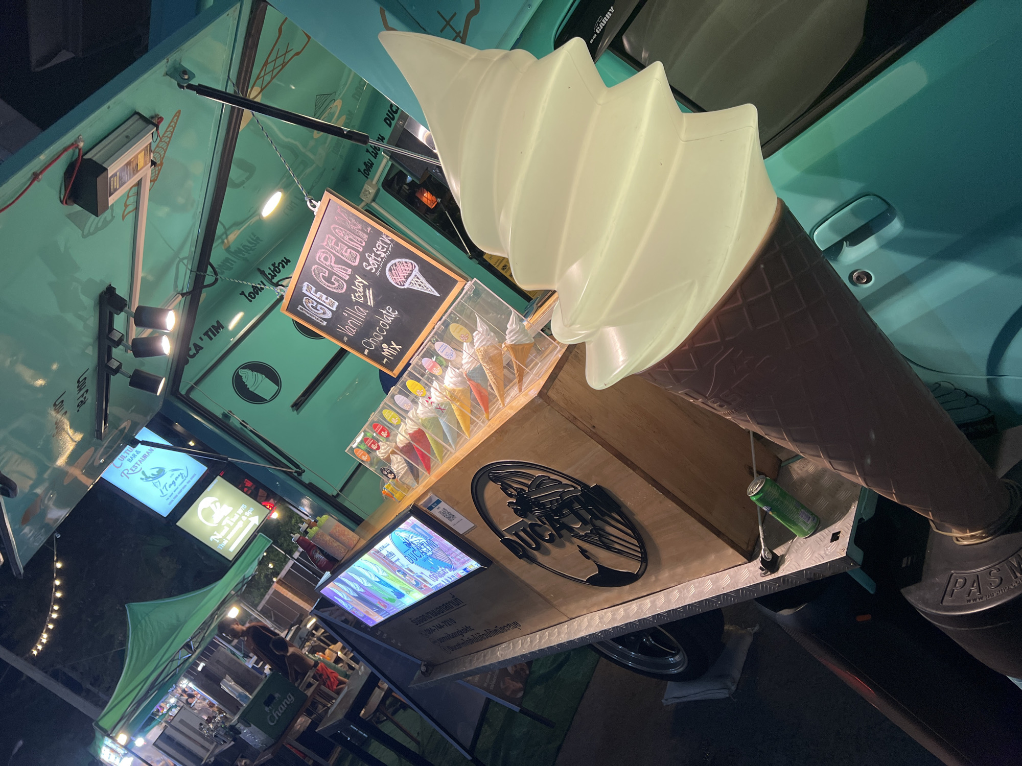 This is a low-angle shot of a food truck, likely at night. The truck is teal and features a large plastic ice cream cone decoration. A chalkboard menu advertises "Ice Cream: Vanilla, Chocolate, Mix," and a display case holds pre-packaged ice cream cones. Signs indicate the truck is located in Baan Bo Phut, Thailand. Visible nearby are signs for "CULTU BAR & RESTAURAN," "Nund Thei 1975 Thai massage & Spa," and another that reads "Tagay." In the background, there are tents, lights, and people at tables, suggesting a food market or outdoor dining area. A can of soda sits at the edge of the service area, with "Chang" branding.