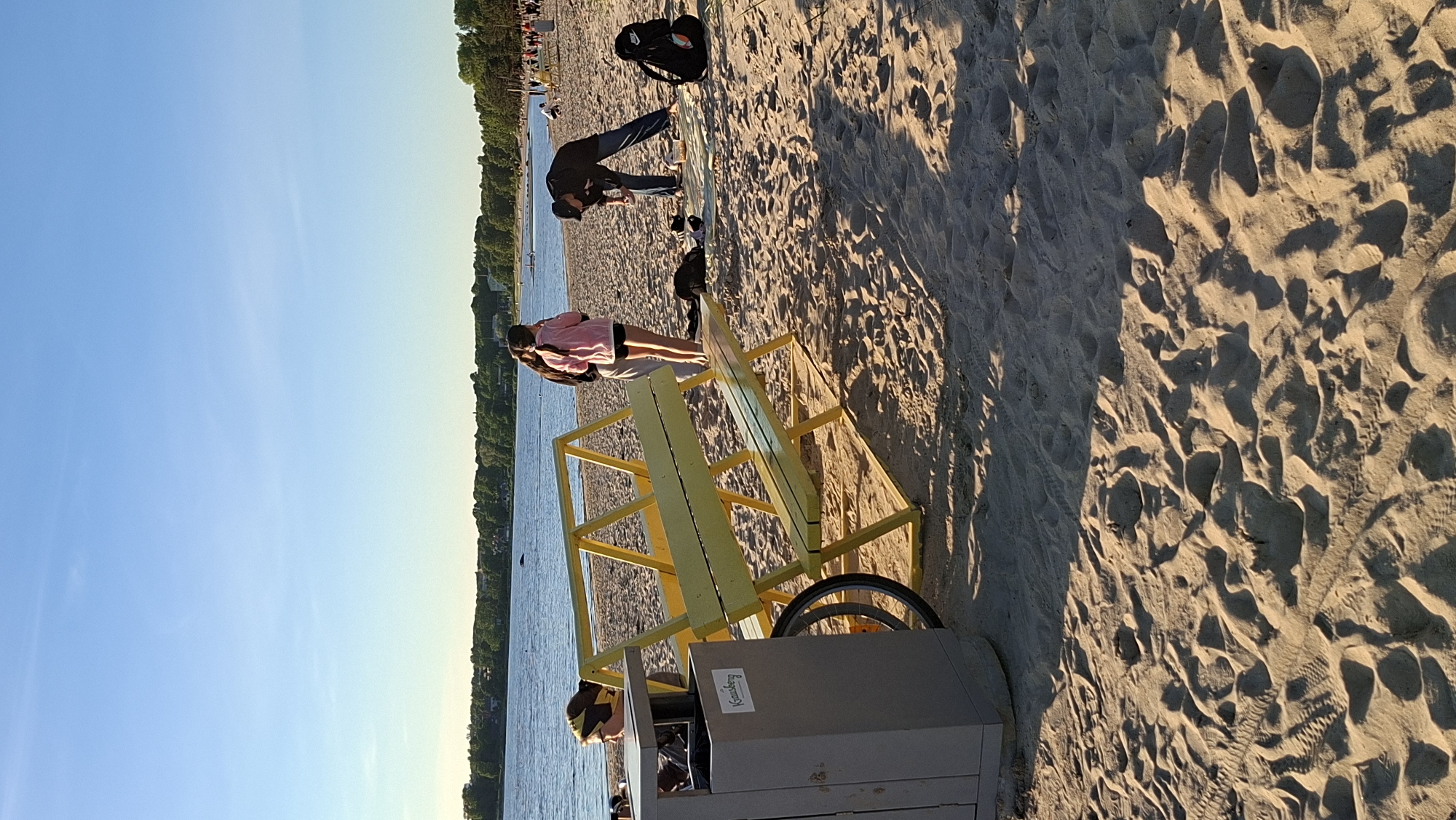 The image depicts a sunny day at a beach, likely in Tallinn, Estonia, given the distinctive yellow bench and a trash receptacle with the inscription "Kansanbergi." The beach is sandy, with rippled textures from the wind and footprints. In the foreground, to the left, is a grey trash bin with a blue bin bag visible inside and a bicycle wheel attached to its base. A bright yellow, modern-looking bench with a geometric backrest is positioned centrally.
Three individuals are visible near the bench. One person, partially obscured, is sitting on the ground or a low surface to the left of the bench, wearing a yellow headscarf and looking away. Two young women are standing near the bench; one in a pink hooded jacket and white shorts, the other in a dark jacket and light-colored pants. They appear to be interacting or observing something. Further to the right, a man wearing a dark jacket, blue jeans, and a cap is bending over on a light-colored cloth spread on the sand, possibly packing or unpacking items. A black backpack with orange accents rests on the sand nearby.
The background shows a calm body of water, likely the sea or a large lake, with a tree-lined coastline in the distance. The sky is clear and blue, indicating fair weather. In the far distance along the coastline, there are hints of other beachgoers and structures. The overall impression is a relaxed, recreational scene at a coastal public space.