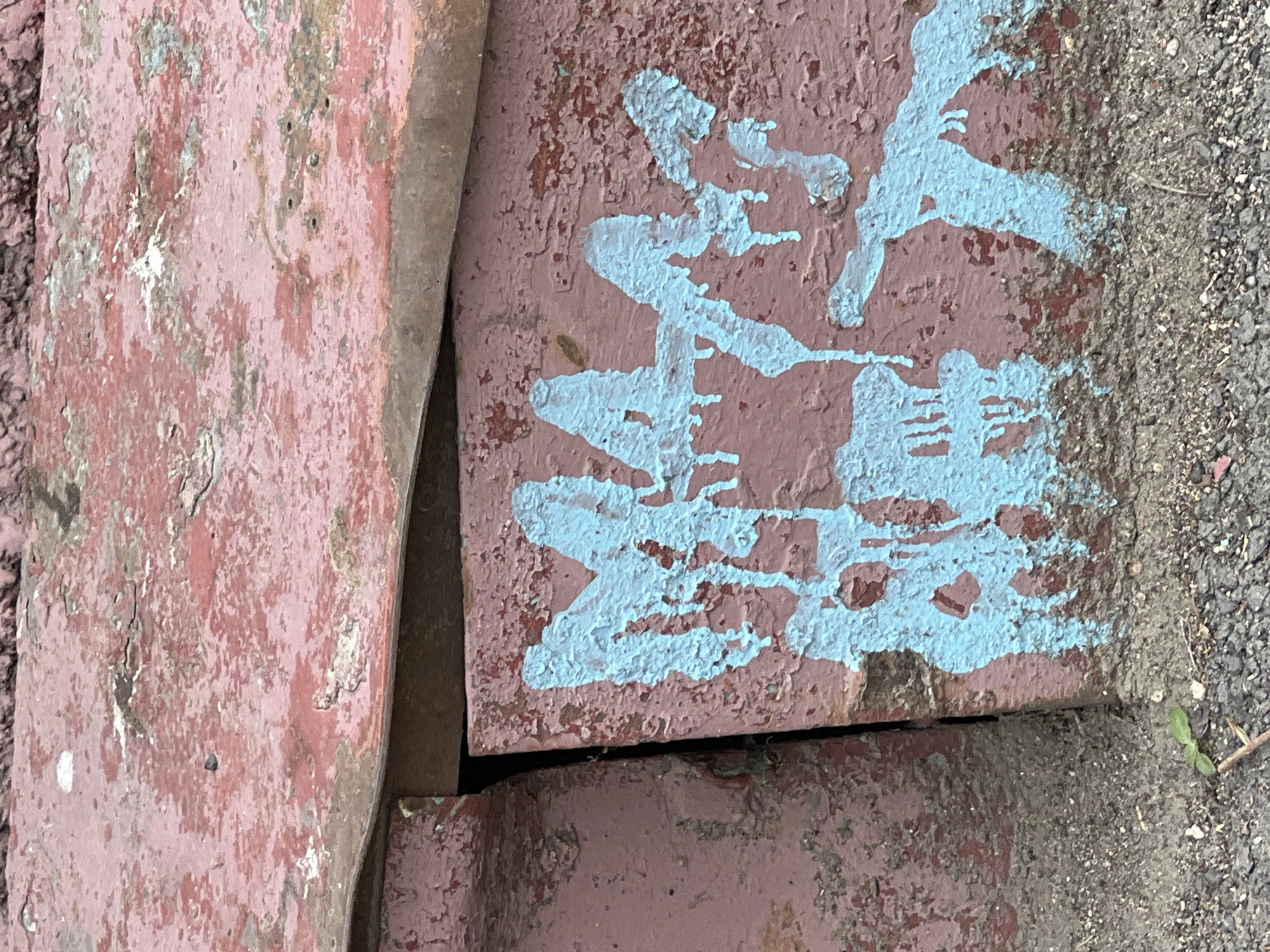 The image shows a close-up view of two weathered, reddish-brown objects, possibly concrete blocks or metal plates, on the ground. One object on the left appears to be a flat slab with peeling paint and a textured surface. The object on the right is a larger block with a similar reddish-brown base color, but it is partially covered with splotches and drips of bright light blue paint. The paint appears to be graffiti or spray paint, applied in a somewhat abstract, gestural manner. The edges of the objects are rough and show signs of age and wear. To the right of the objects, there is a patch of dark, granular material, likely gravel or asphalt, with a small green seedling growing out of it. The lighting suggests it is daytime, and the overall scene appears to be an outdoor, possibly urban or industrial, setting. There is no visible text in the image.