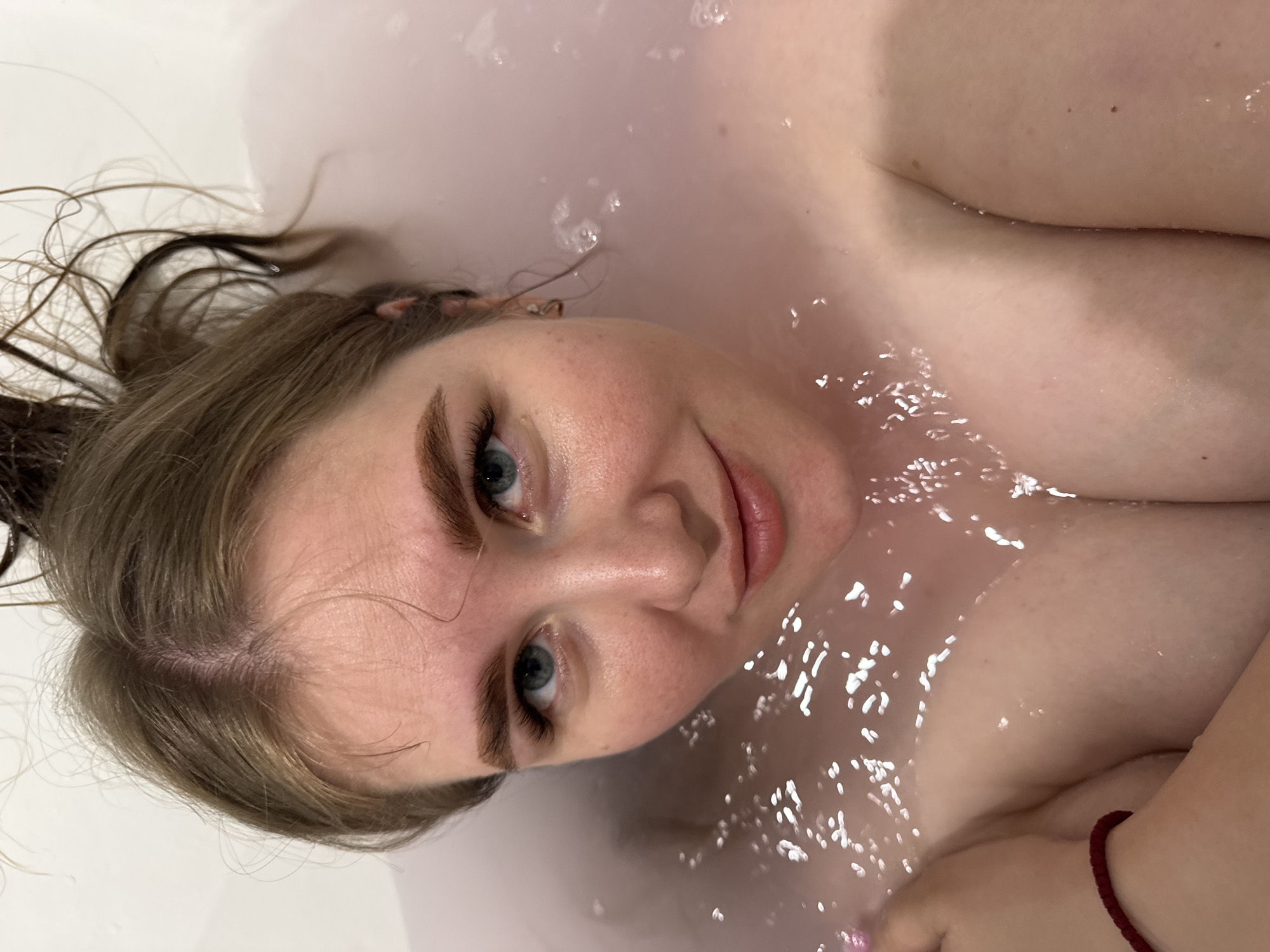 The image displays a woman relaxing in a bathtub, captured from a high-angle overhead perspective. She is lying on her back with her head slightly elevated, looking directly into the camera with soft, blue eyes. Her expression is serene, featuring a gentle smile. Her light brown or dirty blonde hair is wet and floats around her head in the water.
The water in the tub appears milky white, with numerous bubbles and reflections scattering light across its surface. Parts of her upper body, including her shoulders and chest, are visible above the waterline, suggesting partial submersion. A thin, red bracelet is noticeable on her left wrist. Her skin is fair, and her eyebrows are well-defined. The lighting is even and soft, contributing to a calm and intimate atmosphere.
The activity depicted is bathing or relaxing in a bathtub. There are no specific cues for the time of day or weather, but the setting is clearly indoors. No visible text is present in the image. The provided location context for this media file is Ферма, Russia.