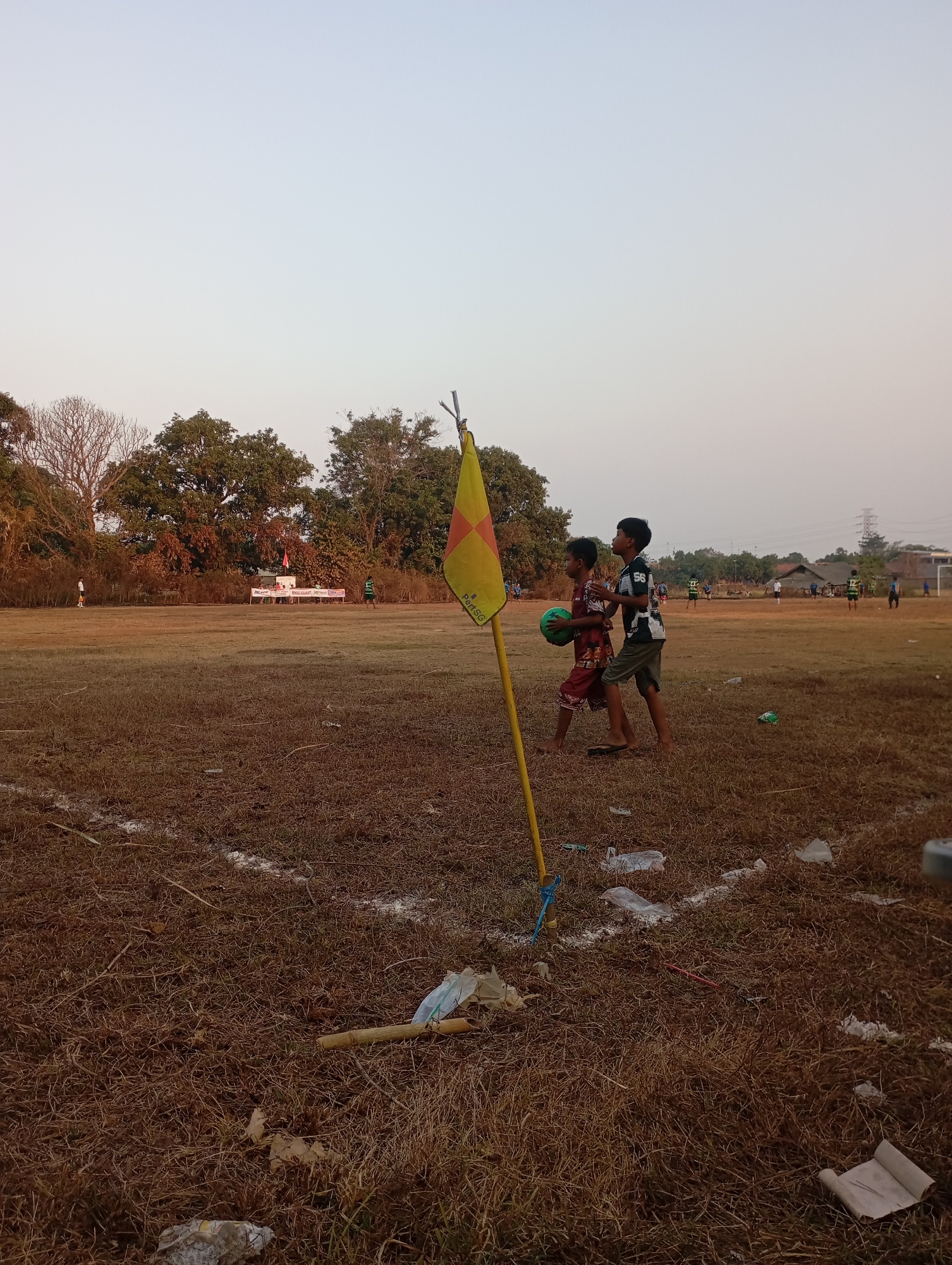 Two young boys are walking on a dry, brown grassy field, possibly a soccer field, in front of a yellow corner flag. One boy is holding a green ball and wearing a red and brown patterned shirt and shorts. The other boy is wearing a black and white patterned shirt with the number 56 on it, and grey shorts. In the background, there are trees and distant structures including what appears to be a grandstand with a banner. Several other people are scattered across the field in the distance, some possibly playing soccer. The sky is a pale, hazy blue, suggesting either early morning or late afternoon. The field is littered with dry grass and some pieces of white and clear plastic debris, as well as a bamboo stick. The scene is set in Kecamatan Haurgeulis, Indonesia.