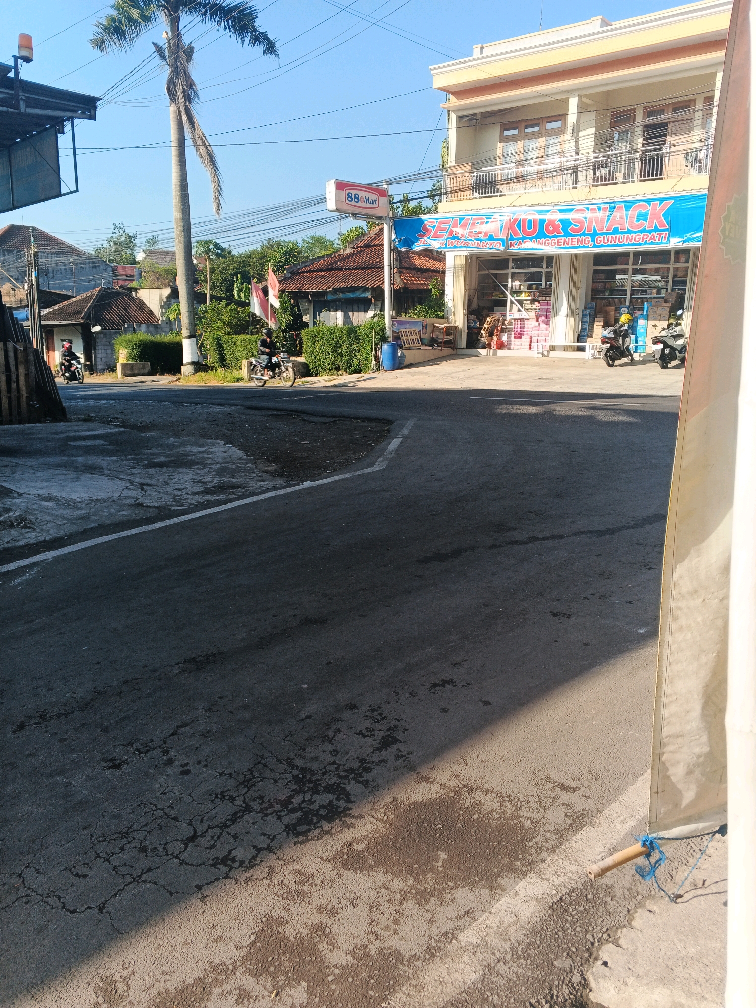 The image depicts a street scene in Gunungpati, Semarang, Indonesia, on a clear, sunny day. In the foreground, a paved road with cracks and patches is visible, with a white line marking the lane. The mid-ground features a "Sembako & Snack" store with a large blue banner displaying the name and address. Two motorcycles are parked in front of the store, one black and one white. A person on a black motorcycle is riding away from the camera. Further back, a tall palm tree stands prominently, with a 88 Mart sign positioned to its right. Several residential buildings with traditional tiled roofs are visible behind the store and palm tree. Two Indonesian flags are seen flying from a pole near the store. In the background, lush green bushes and trees form a backdrop under a clear blue sky. The overall impression is one of a typical neighborhood commercial area.
