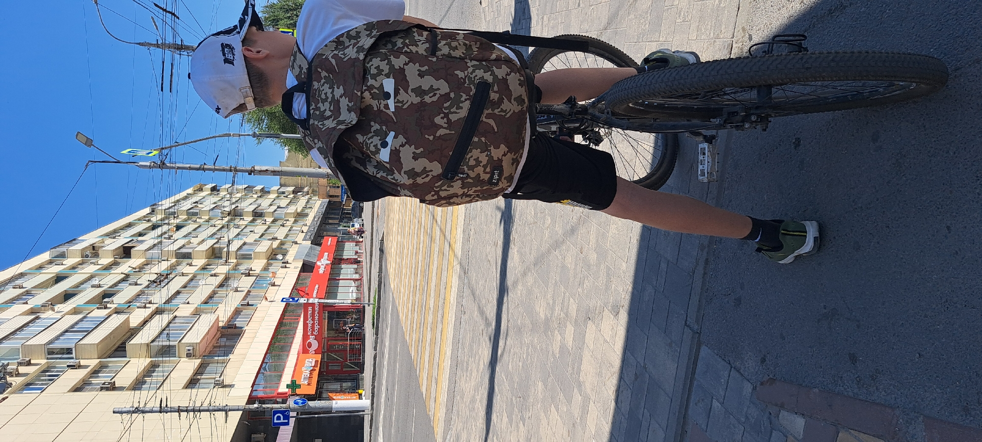 A young person wearing a white baseball cap and a camouflage backpack is sitting on a bicycle on a sunny day. The person is wearing black shorts and green athletic shoes. The bicycle is a dark color with knobby tires. The scene is set in an urban environment with a large apartment building in the background. In front of the building, there are several storefronts, including one with a red awning. A pedestrian crossing with yellow and white stripes is visible on the street. Power lines and streetlights are also present in the scene. The ground is paved with concrete and asphalt.