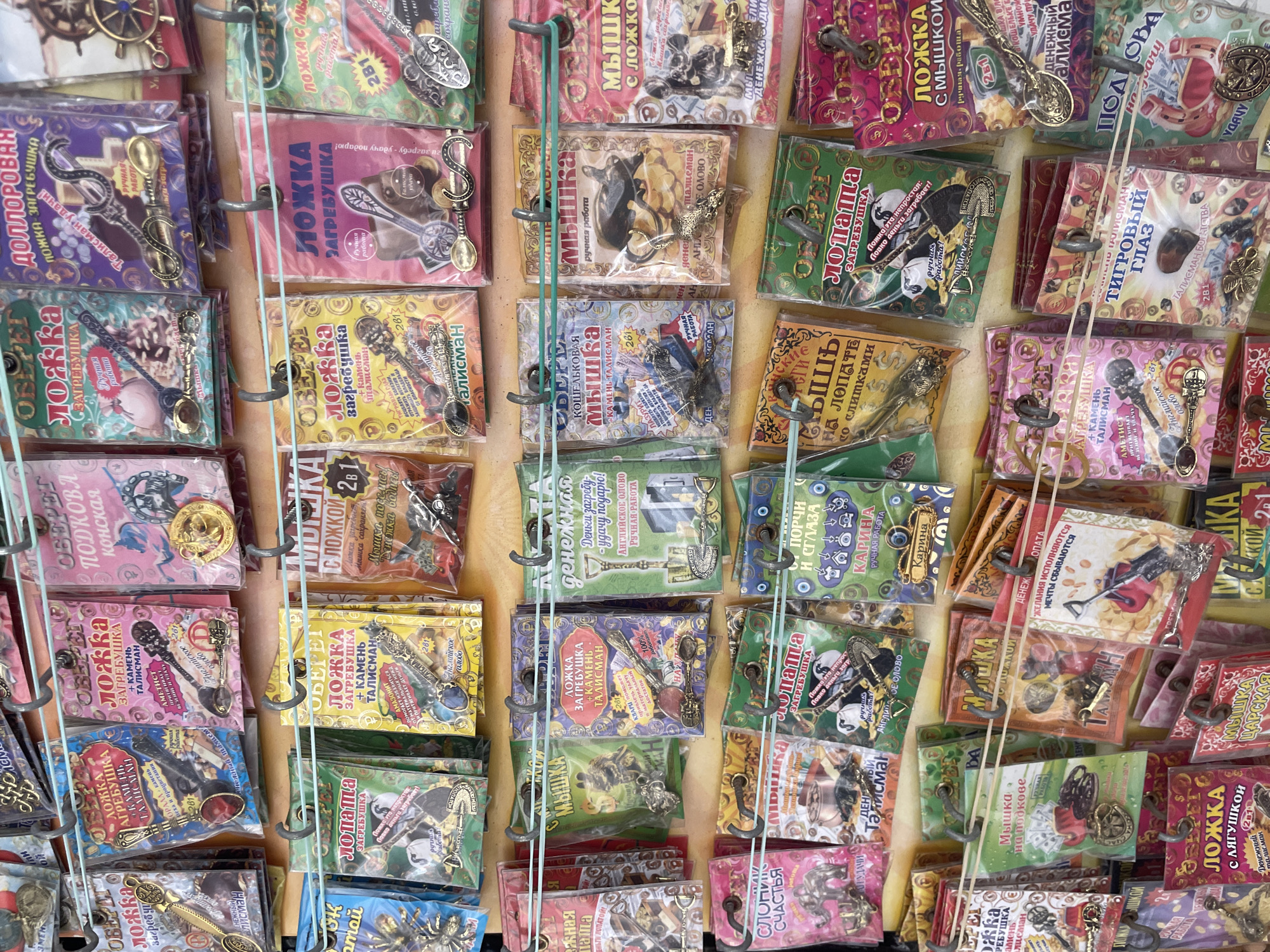 The image depicts a close-up view of a display of souvenir items. These souvenirs are small, packaged metal spoons or shovels attached to cards printed with text in Russian. The cards vary in color and design, featuring images of animals (mice, frogs), coins, or symbols of wealth. The spoons and shovels are metallic and have decorative designs.
The items are arranged in a grid-like pattern and are suspended from a metal rack. A translucent green elastic cord is visible, used to secure the items to the rack. The background is a light brown or beige color.
The image suggests a souvenir shop or market stall in Olginka, Russia, where tourists may purchase these items as reminders of their visit. The scene appears to be a commercial setting.