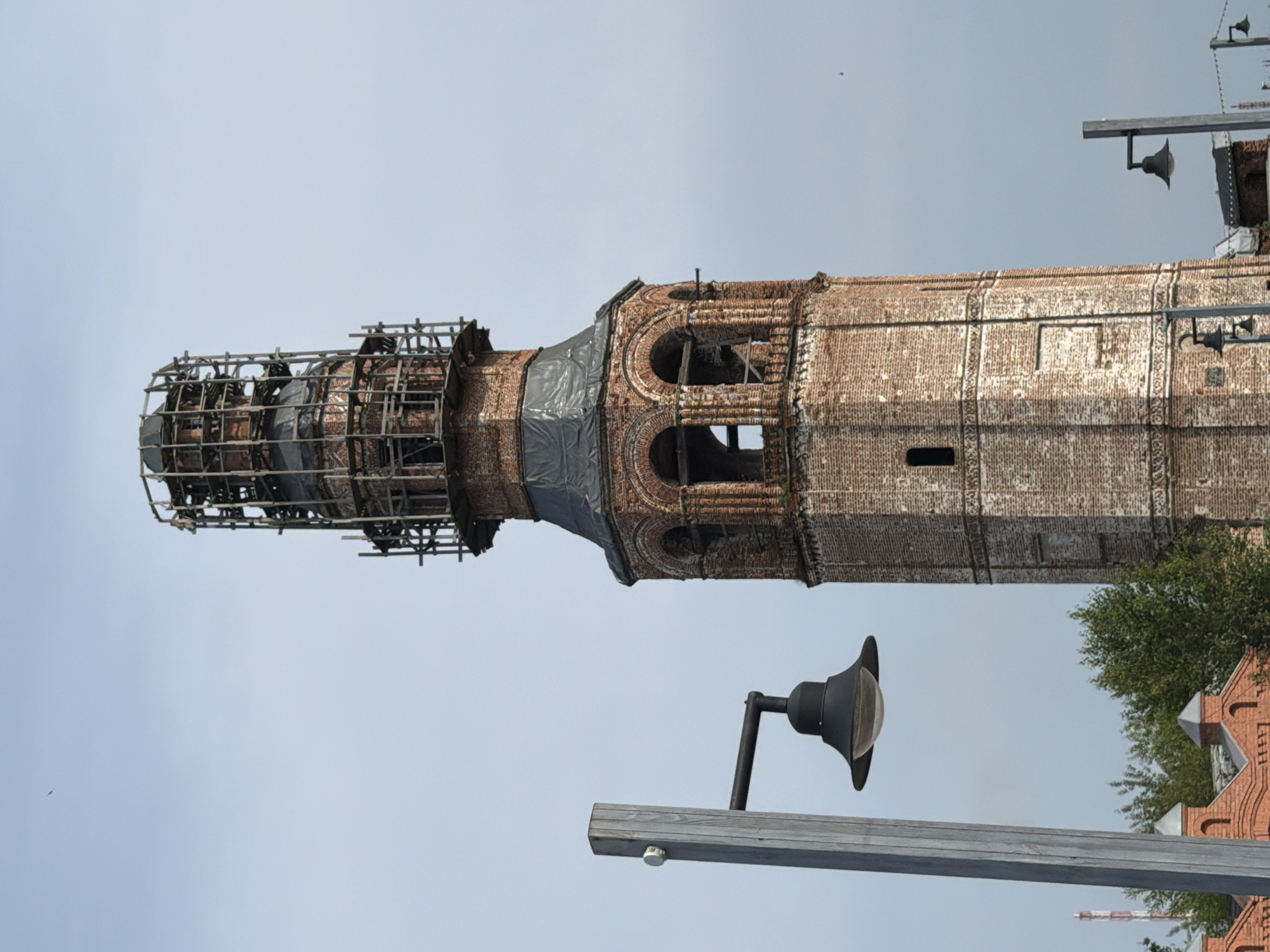 A brick bell tower stands tall against a pale sky. The tower is octagonal in shape and made of weathered red brick, with decorative bands of lighter brickwork. The top of the tower is covered in scaffolding, suggesting ongoing repairs or restoration. Several arched openings are visible in the upper section of the tower. In the foreground, a modern lamppost with a black lamp stands to the left of the tower. Further to the left and below, parts of other brick buildings with pointed roofs are visible, along with green trees. Another lamppost is visible in the lower right quadrant of the image. The scene appears to be in the city of Usolye, Russia, with the bell tower likely being a prominent landmark.