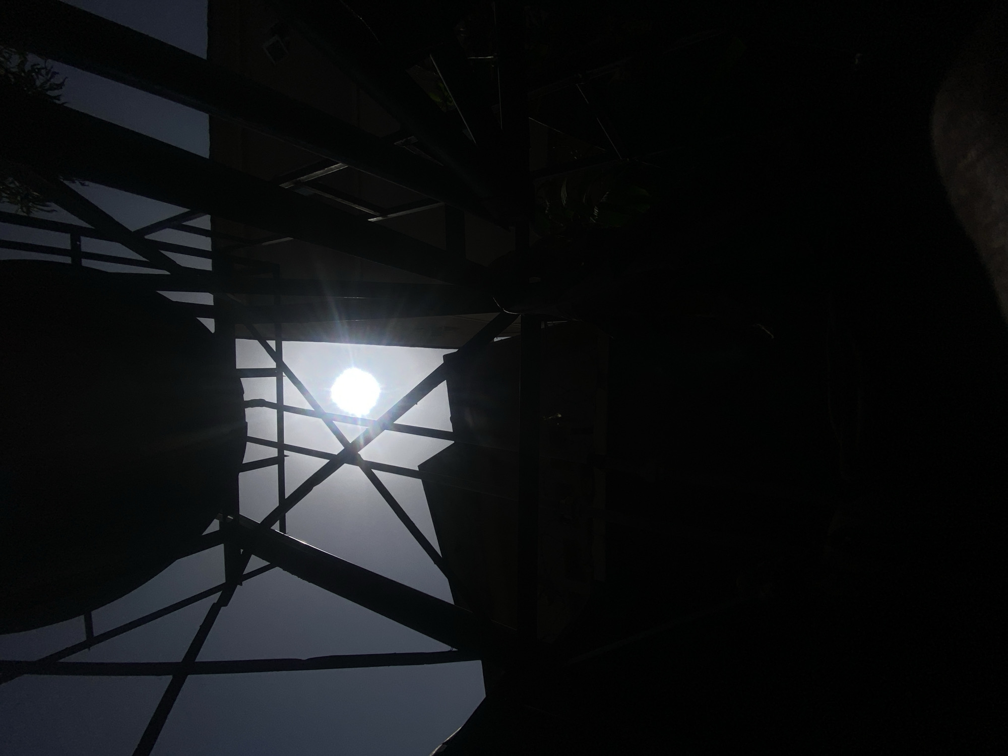 The image captures a heavily backlit outdoor scene, likely located in Barden Yana, Nigeria, dominated by a stark contrast between a brilliant light source and dark, silhouetted structures. At the center-top of the frame, a powerful, circular light, presumably the sun, shines intensely, creating a sunburst effect. This light strongly illuminates the sky directly around it, rendering it a lighter gray.
Surrounding and partially obscuring the light are numerous dark, angular forms. These appear to be the structural framework of a building, scaffolding, or a robust overhead covering, composed of intersecting beams, rods, and possibly corrugated sections. These elements are rendered in deep silhouette, almost entirely black due to the extreme backlighting, showing virtually no internal detail or texture. Some areas on the left and right sides of the image are plunged into complete darkness, suggesting large objects or structures casting vast shadows.
The dramatic lighting indicates a clear, sunny daytime, possibly midday or early afternoon, given the high position of the light source. No specific activity, people, or discernible objects other than the framework are visible. There is no visible text. The image's composition emphasizes geometric forms and strong contrasts.
