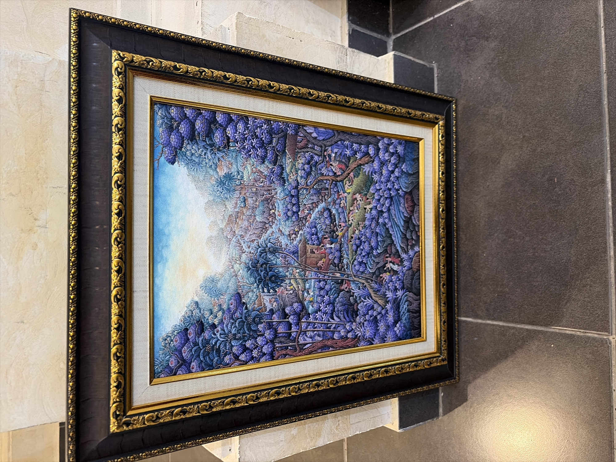 A framed painting depicts a dense, stylized Balinese village scene rendered in vibrant purples, blues, and greens. The artwork is leaning against a textured cream-colored wall with a black tiled floor below. The painting is framed by a thick, dark wood frame with ornate gold detailing.
The scene within the painting is a panorama of lush vegetation, with rolling hills and dense foliage dominating the landscape. Small, traditional Balinese houses with thatched roofs are nestled amongst the trees and bushes. Numerous small figures, likely villagers, are scattered throughout the scene, engaged in various activities such as tending to crops, carrying baskets, and interacting with each other. The sky is a soft blue with hints of yellow, suggesting either sunrise or sunset. The overall impression is one of a peaceful, communal village life set within a tropical environment. The style of the painting is reminiscent of traditional Balinese art, known for its intricate detail and vibrant colors.