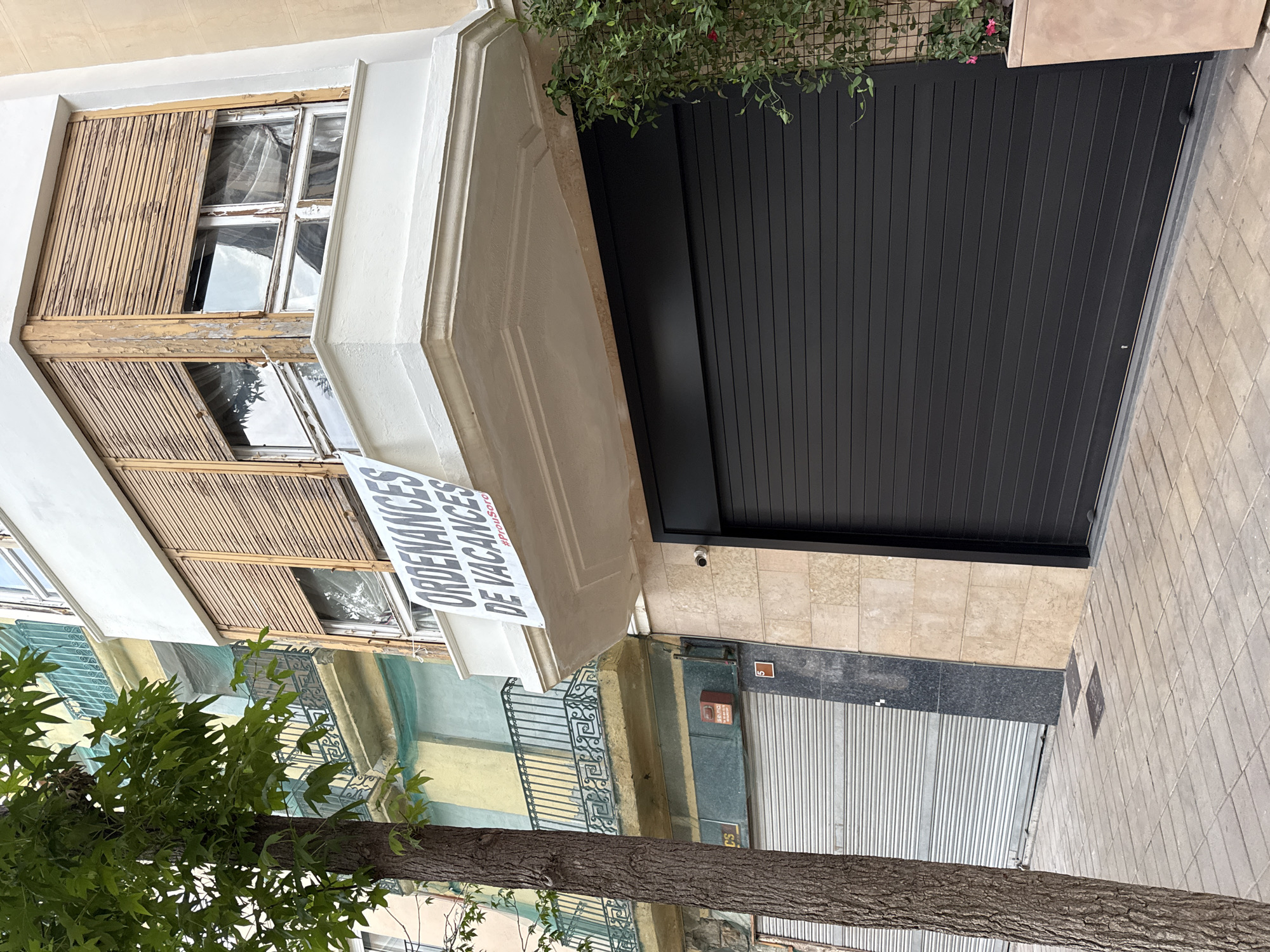 The image shows a street scene in the city of Igualada, Spain. In the foreground, a large black roller shutter door is visible, covering the ground floor of a building. To the left of the roller shutter is a light-colored tiled wall, and to the left of that, a silver roller shutter door. A mature tree with green leaves stands on the sidewalk to the left. Above the black roller shutter, a white balcony with weathered wooden shutters is visible. A banner with text hangs from the balcony, reading "ORDENANCES DE VACANCES" and "#ProuSor". Above this balcony, another building with a green-painted balcony and yellow and light blue painted walls is partially visible. The environment appears to be urban, with paved sidewalks and buildings lining the street.