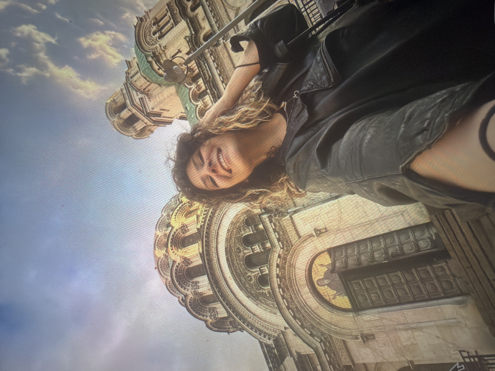 The image depicts a low-angle selfie of a young woman smiling brightly in front of a grand, multi-domed Orthodox cathedral, located in the city of Shumen, Bulgaria.
The woman, with shoulder-length curly hair featuring brown and blonde highlights, occupies the lower center-right of the frame. She wears a black leather jacket over a dark top and a thin silver necklace with a small pendant. Her head is tilted slightly back and to the right, revealing a wide smile, and her right hand is raised, gently touching the hair on the right side of her head. A dark band or bracelet is visible on her left forearm.
The architectural background is dominated by a large, ornate cathedral. Its most prominent features include a broad central dome on the left, adorned with tiered arches and windows, and a taller, narrower bell tower with a smaller dome on the right. Both domes exhibit a distinct green patination. The cathedral's façade is constructed from light-colored stone, featuring elaborate carvings and multiple arched openings. Directly behind the woman's left shoulder, above a pair of dark, slightly ajar double wooden doors, an arched mosaic depicts a robed figure with outstretched arms. The sky above is light blue with scattered white and warm-toned clouds. The overall lighting suggests an outdoor scene under natural light, possibly an overcast day or during twilight, imparting a warm glow to the stone façade.