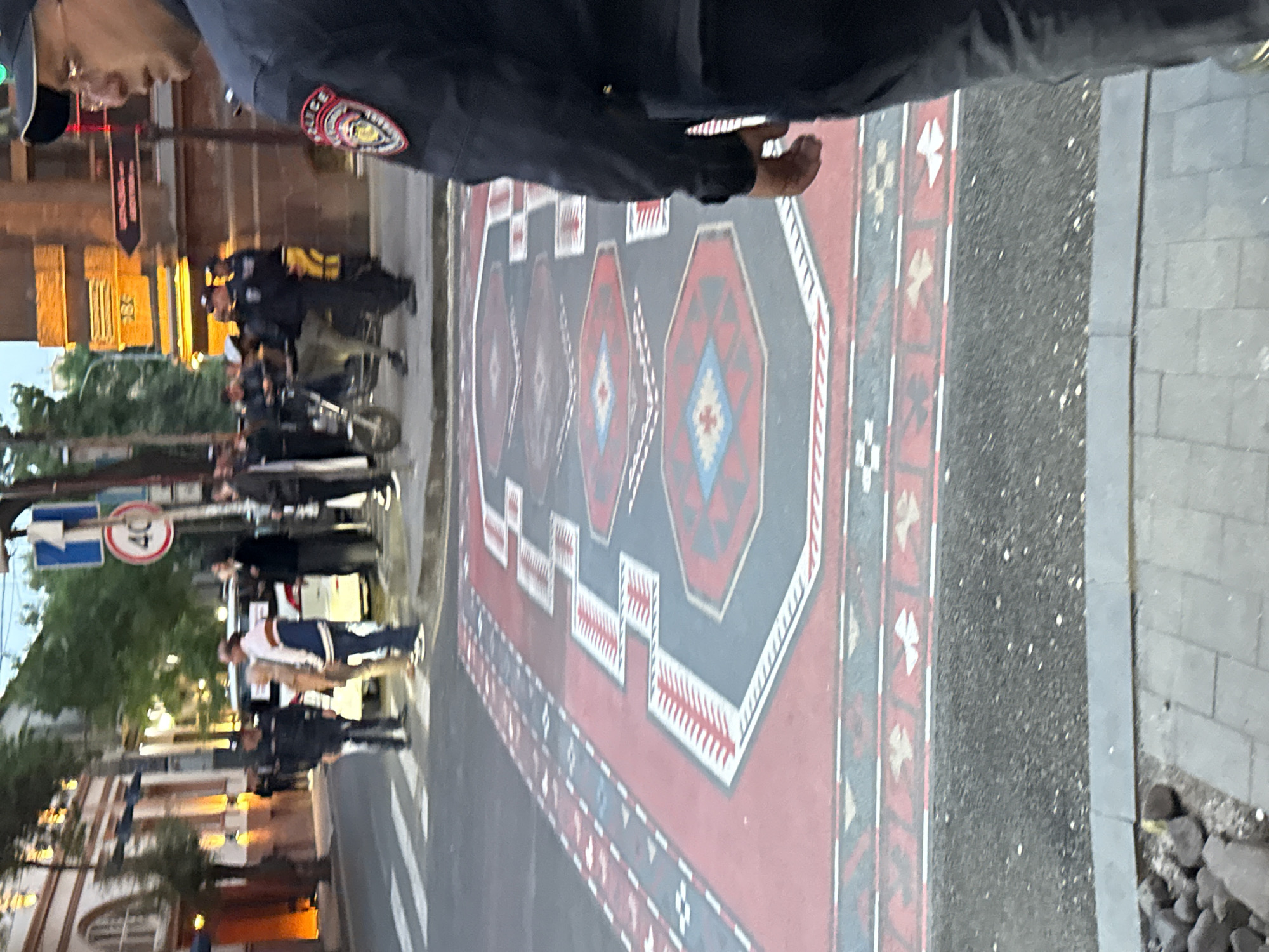 The image shows a street scene in Yerevan, Armenia, featuring a large carpet design painted on the road surface. A group of people are seen walking, some accompanied by police officers in uniform. A speed limit sign of 40 km/h is also visible. Buildings line the street, and there are trees and streetlights present, indicating an urban environment. The image appears to have been taken from a slightly elevated position.