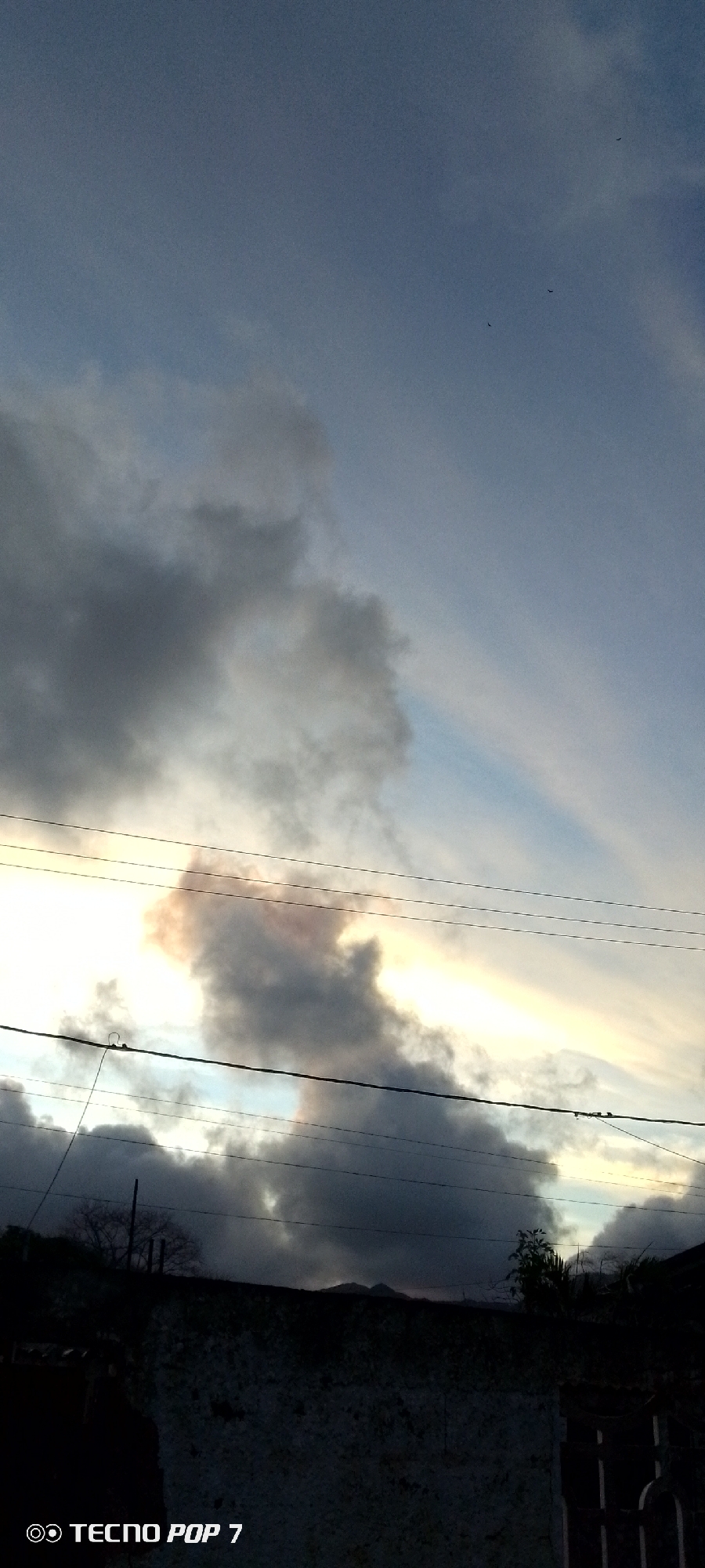 This image captures a dramatic sky scene in Altagracia, Venezuela, likely during the late afternoon or early evening given the quality of light. The primary subjects are the clouds and sky, with foreground elements including power lines and dark building structures.
The sky is a mix of heavy, dark grey clouds and patches of lighter blue. A significant portion of the image's center is dominated by a large, dense cloud formation. Behind and through these clouds, a bright, warm light, ranging from yellow to orange and reddish hues, creates a striking glow, suggesting either sunrise or sunset. This light illuminates parts of the clouds from behind, highlighting their contours and textures.
Several power lines traverse the frame horizontally, silhouetted against the bright sky and darker clouds. In the upper right quadrant, against a clearer blue sky, three small, dark specks are visible, appearing to be birds in flight.
The lower section of the image is dark and silhouetted. It shows the top edge of a building or wall, made of what appears to be rough concrete or stone. Sparse, dark foliage and possibly distant hills or mountains are visible just above this structure, beneath the cloud line.
The overall mood is one of natural drama and beauty, with the contrast between the vibrant light and the heavy clouds. The text "TECNO POP 7" is present as a watermark in the bottom left corner of the image.