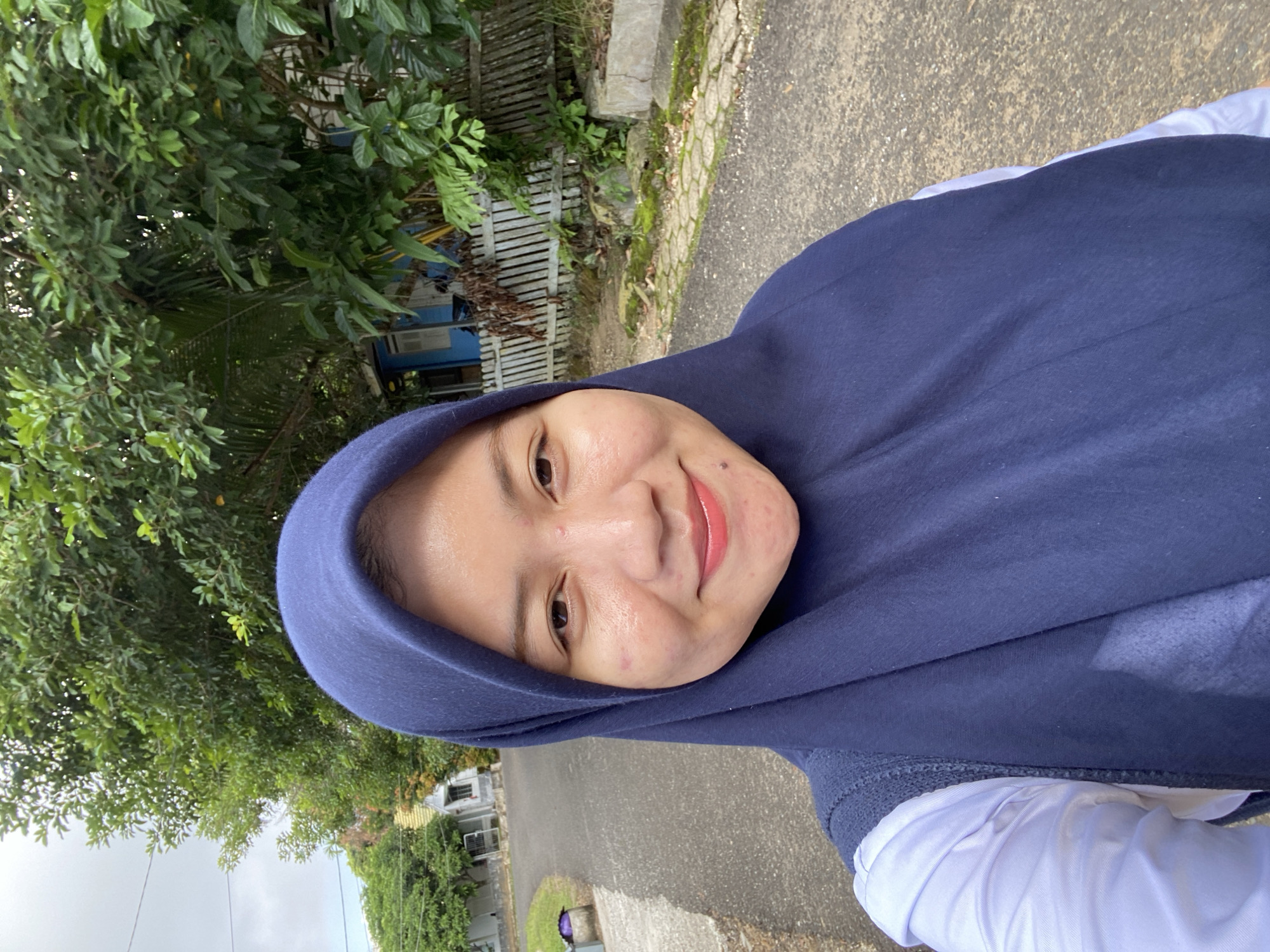 A selfie shows a woman wearing a navy blue hijab and a white shirt. She is standing outdoors on a paved road, with trees and houses in the background. The area appears to be a residential street in the city of Banjarbaru, Indonesia. The woman is smiling directly at the camera, and her face is well-lit. She has some blemishes on her skin. The sky is overcast. To the right of the woman, a wooden fence and lush green vegetation are visible. A small blue building can be seen behind the fence. The road is gray asphalt, with a lighter colored shoulder on the left side. A small trash receptacle is present on the shoulder. The overall impression is a casual outdoor portrait.