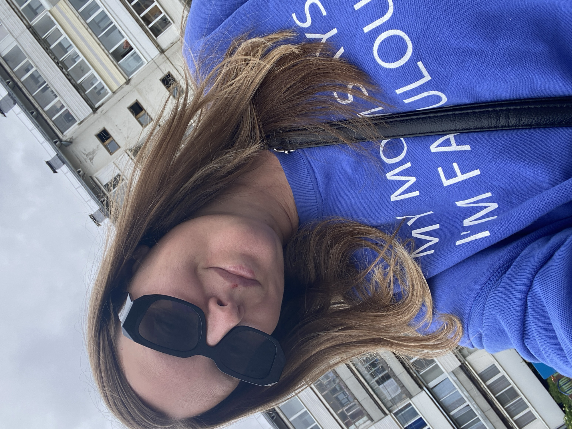 A young woman with long, light brown hair and black sunglasses takes a selfie outdoors. She is wearing a blue sweatshirt with white lettering that reads "MY MOM SAYS I'M FABULOUS." She is positioned in the foreground, with a tall, multi-story apartment building in the background. The building appears to be made of concrete and has many windows. The sky is overcast. The location is Noviy, Russia.