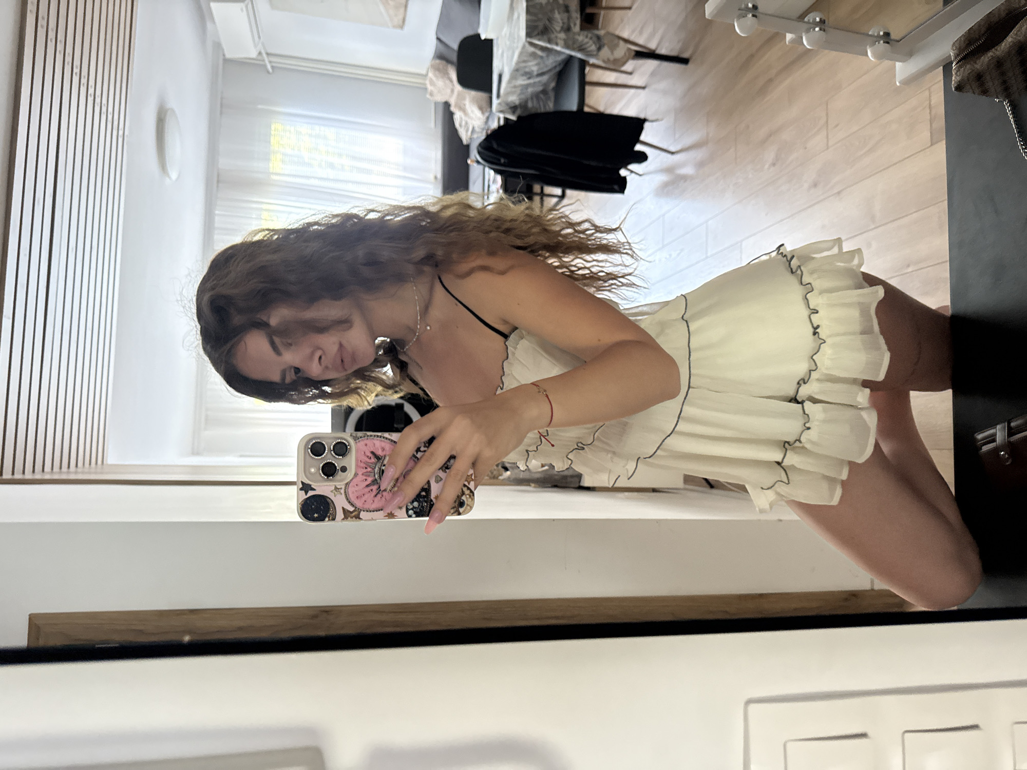 A woman is taking a selfie in a room. She is wearing a light-colored dress with frills and has long, curly hair. She is holding a phone with a decorated case. The room has a mirror and what appears to be furniture, including chairs and a coat. There is also a window visible. The location is not specified.