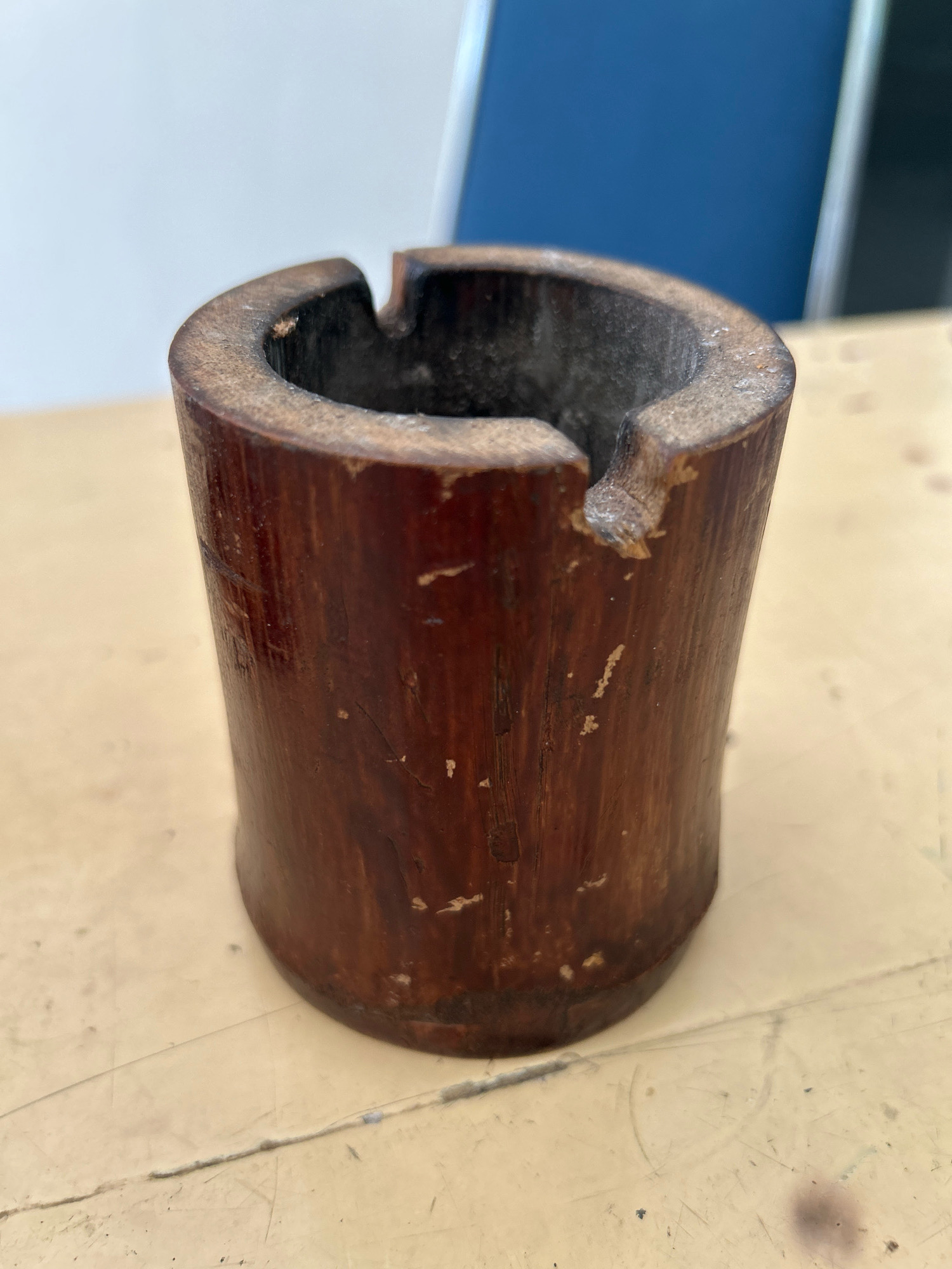A close-up, eye-level shot features a hollowed-out piece of dark brown, weathered wood, likely a section of bamboo, positioned on a beige, textured surface. The wood has a cylindrical form with an open top and a noticeable notch carved into its upper rim, suggesting it might be an ashtray or a rustic holder. The surface of the wood exhibits a grain pattern and some minor abrasions and chipped areas. The background is blurred, with a light-colored vertical plane on the left and a dark blue vertical plane on the right, indicating an indoor setting. The lighting appears to be natural or overhead, casting soft shadows and highlighting the texture of the wood and the surface it rests on. There are no people or visible actions occurring in the scene. The image appears to be a still-life shot of a single object. This object is located in an unidentified indoor environment within the city of Sabang, Indonesia.