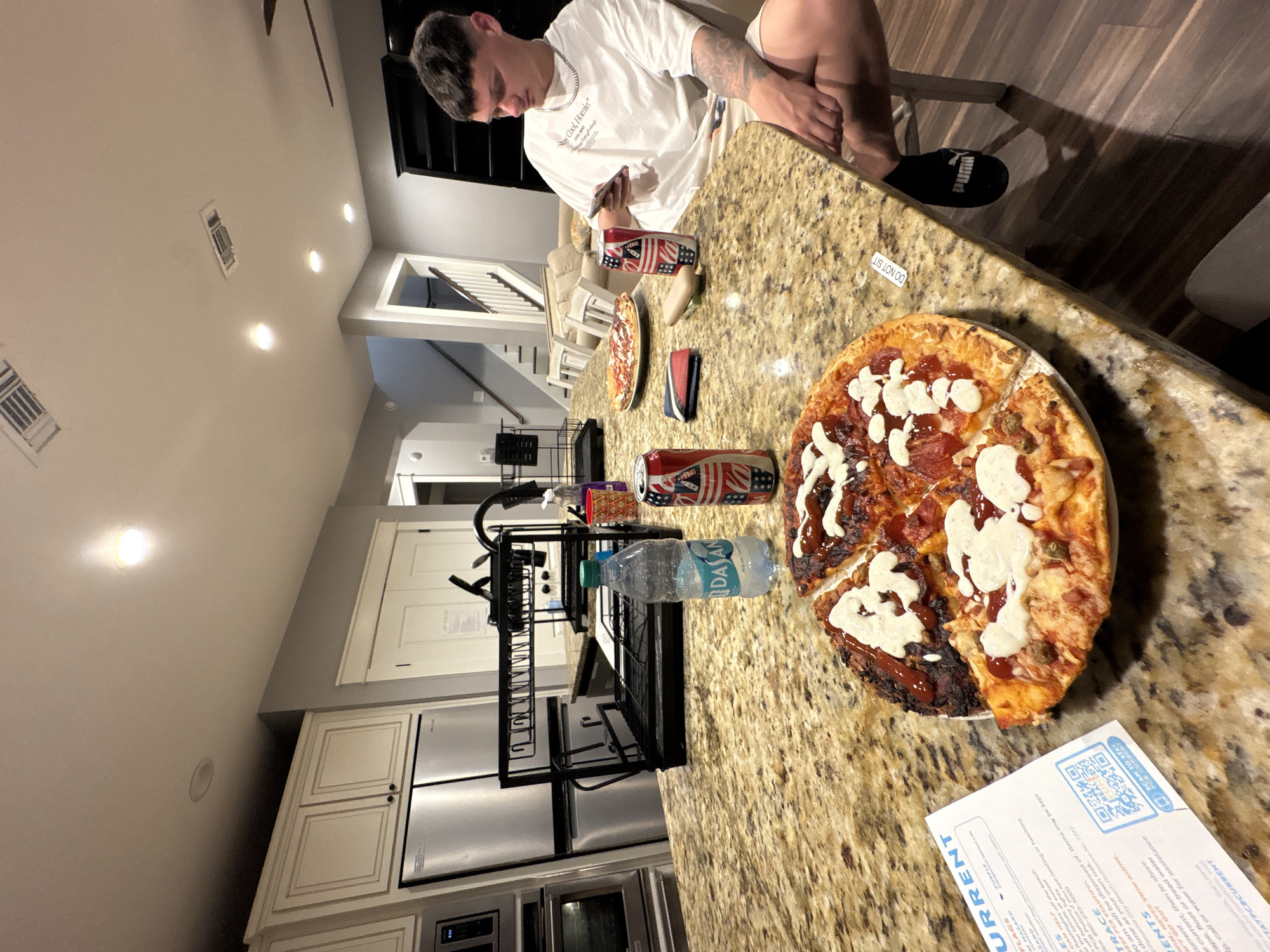 Here is a description of the image.
A person sits at a counter in a home kitchen in Panama City Beach, Florida. The person is white, with dark hair, wearing a white t-shirt with the phrase "Stay Cool, Homie" and shorts. They are holding a phone in their hands and looking at the phone. The counter is granite and has a pizza, cans of soda, and a bottle of water on it. There is a receipt on the counter as well. The receipt has the word "CURRENT" on it. The receipt says "Stay PCB Current", and it talks about trace elements and other information about Panama City Beach, Florida. There is a drying rack and kitchen appliances to the left of the pizza. The floors are wood. There is a staircase visible in the background. A sticker on the counter says "DO NOT SIT".