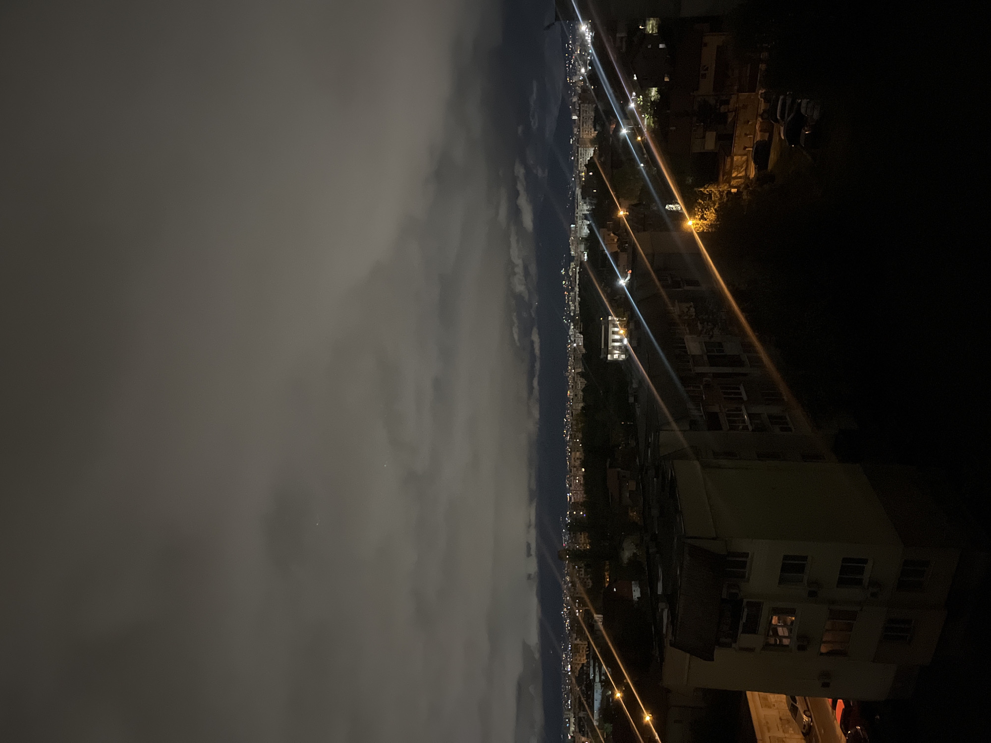 The image captures a panoramic view of a city at night, likely Sofia, Bulgaria, from an elevated vantage point. The upper half of the scene is dominated by a heavily clouded, dark sky, displaying dense grey cloud formations that subtly reflect the distant city lights.
Below the distinct cloud line, the urban landscape extends across the frame, characterized by a multitude of artificial lights. These lights, appearing as bright pinpoints and distinct streaks of white, yellow, and orange, delineate streets and buildings, illustrating the city's spread. These prominent light streaks suggest the presence of bright street lamps or urban infrastructure.
Numerous buildings are visible, forming the urban tapestry. In the foreground to the right, a large building, possibly residential, is partially illuminated, with some windows glowing amidst the general darkness of its facade. Further into the midground, the lights intensify, indicating a denser cluster of city structures.
No people are visible within the frame, and there is no discernible text. The overall atmosphere conveys the quiet, expansive nature of a city illuminated after dark under a cloudy sky.
