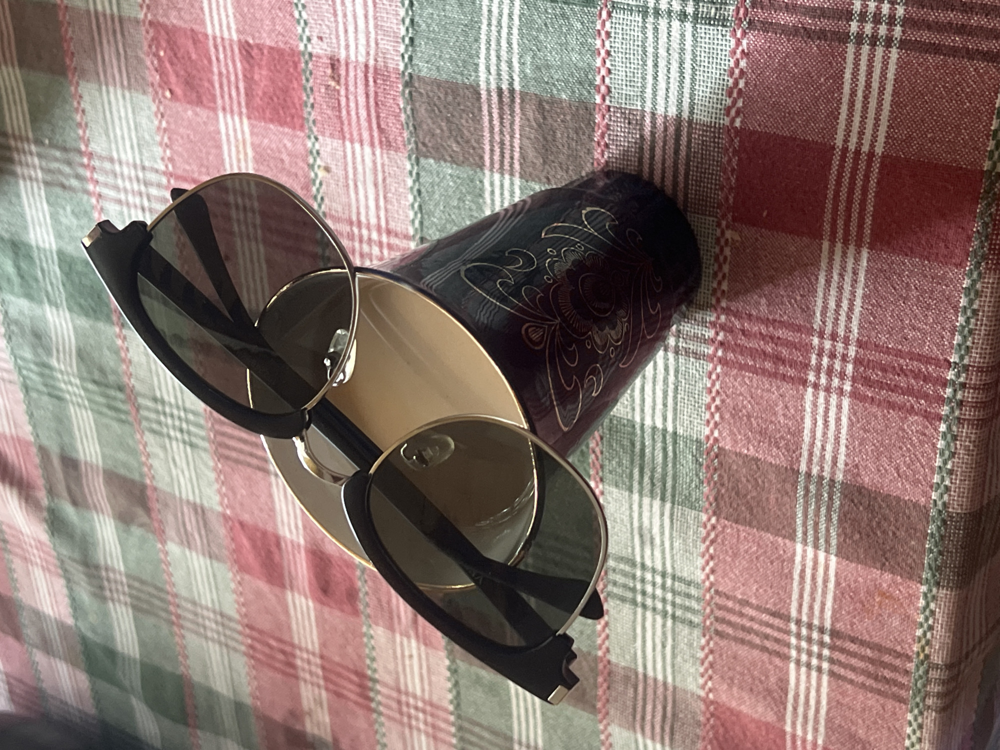 A pair of black-rimmed sunglasses rests on a decorative dark purple mug, which is itself placed on a red and green plaid tablecloth. The scene is likely set indoors, given the nature of the objects and the tablecloth. There is no indication of the location being in the city of Norviliškės, Lithuania. The lighting suggests it is daytime, with the light source coming from above and to the left. There are no people or animals visible in the image.