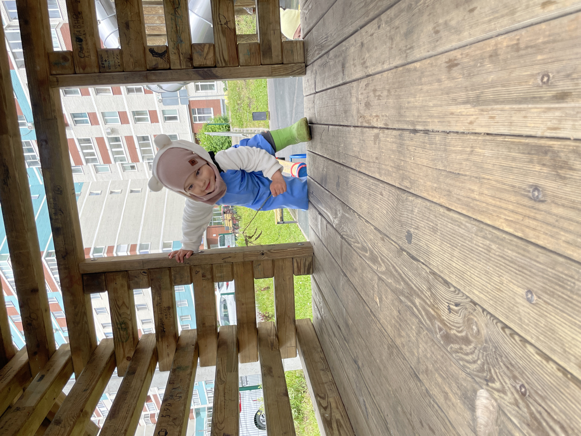 A young child, appearing to be a toddler or preschooler, is playfully peeking out from within a rustic wooden play structure on an outdoor playground. The child looks directly at the camera with a gentle smile, their face framed by a light pink hat with two round, ear-like pom-poms. They are dressed in a white long-sleeved top and blue waterproof overalls, with green rubber boots visible on their feet, suggesting cool or damp weather.
The wooden structure is constructed from rough-hewn planks and beams, exhibiting visible grain and some wear. Several beams feature handwritten graffiti or carved markings. Legible text includes "САША ТАВАН", "МАРИНА", and numerical sequences like "1984" and "27 06 91".
In the background, behind the play structure, multi-story residential apartment buildings with numerous windows are visible, typical of a city environment. Green grass and some foliage are also present, indicating a park or common area. A light-colored vehicle can be partially seen in the distance. The scene is set outdoors during the daytime, likely under an overcast or partly cloudy sky, capturing a joyful moment of play in Izhevsk, Russia.