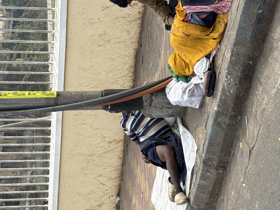 A person wearing a striped shirt and dark bottoms sits on a white cloth near a curb, resting against a utility pole. Behind them, a collection of clothing and personal belongings are piled. The setting appears to be an urban sidewalk, with a building and barred window visible in the background. The overall scene depicts a person living in impoverished conditions, possibly homeless, seeking rest on the street.