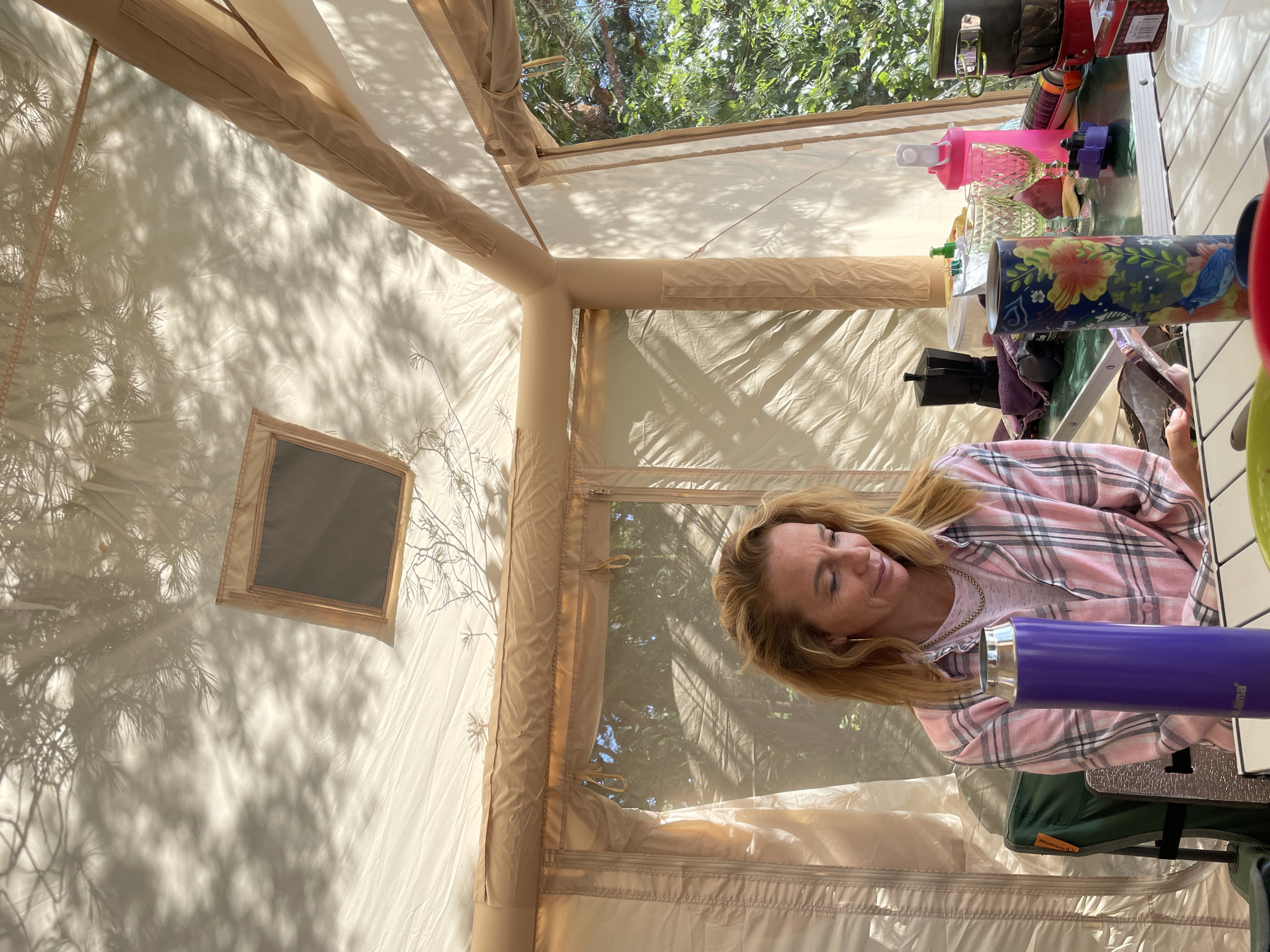 An adult woman with blonde hair and a pink and white plaid shirt sits at a table inside a tan-colored tent. She is looking down at a mobile phone in her hands. On the table in front of her are several items, including a purple water bottle, a patterned cylindrical container, a black coffee maker, two clear wine glasses, a pink shaker bottle, and stacked camping pots. The tent has a mesh window showing trees and foliage outside, and the ceiling of the tent displays shadows of pine needles. The scene appears to be a campsite, possibly in Zelenaya Roshcha, Russia, given the natural outdoor setting.