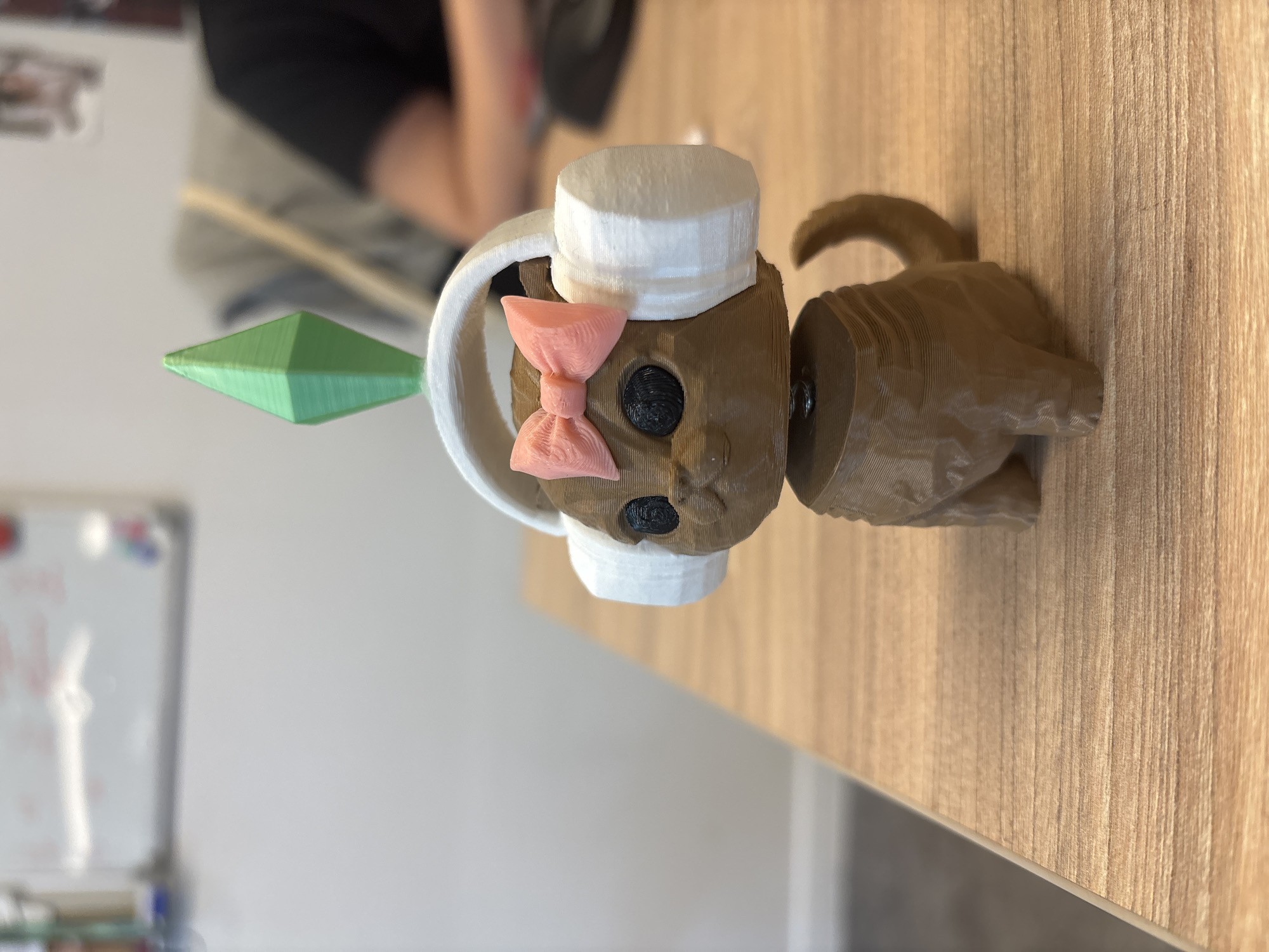A 3D-printed figurine of a cat with headphones and a pink bow is placed on a wooden desk. The cat has black eyes and a green diamond-shaped object on its head, resembling a Sim's Plumbob. The figurine is brown, with white headphones and a pink bow. In the background, a whiteboard with writing and a person wearing a black shirt are visible. The scene is indoors, with a bright, neutral-colored wall and a hint of other office furniture. No specific location in Vladivostok, Russia, can be determined from the image.