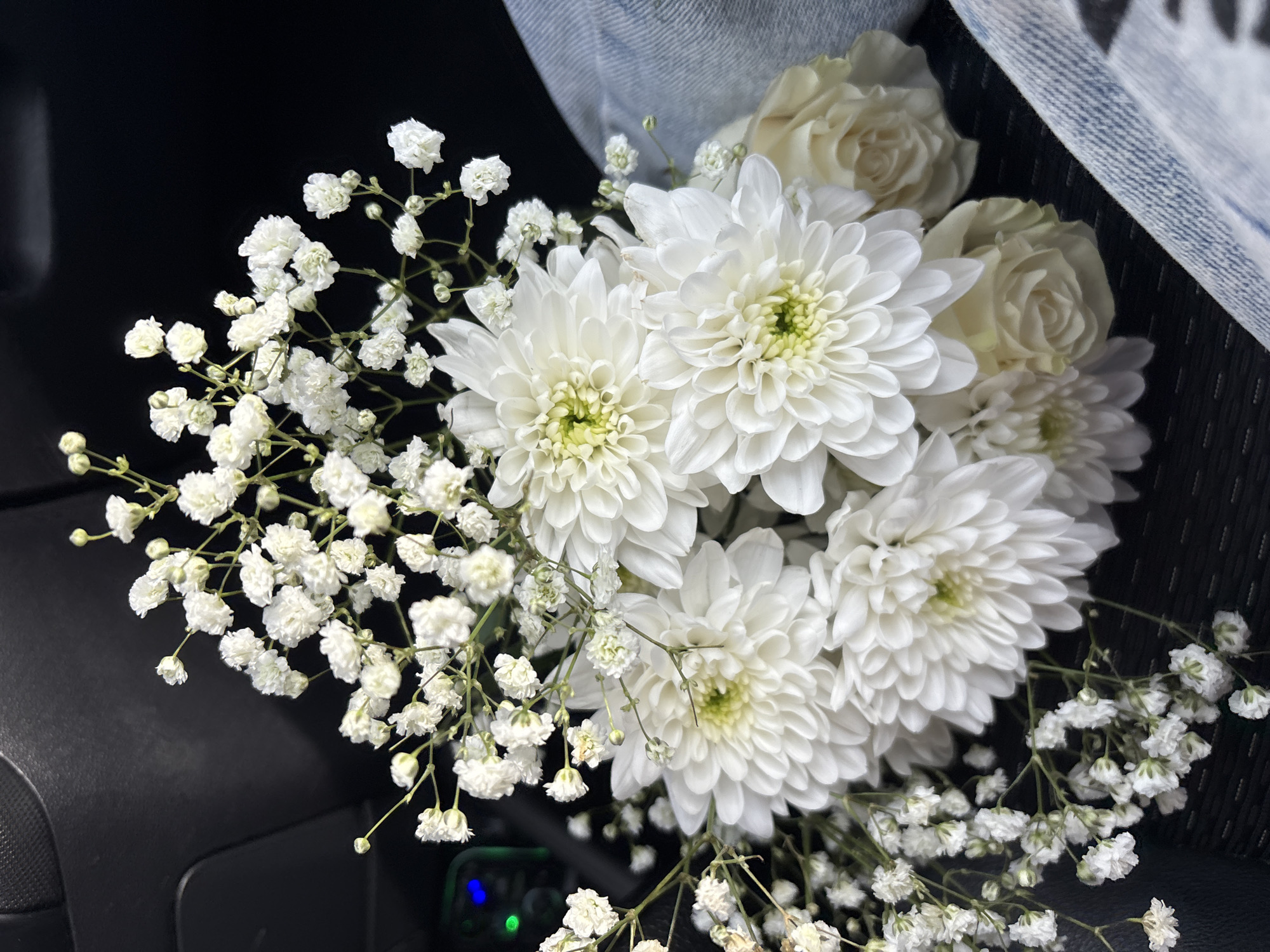 A bouquet of white flowers sits on a dark surface, with a piece of denim fabric partially visible at the top. The bouquet is composed of white chrysanthemums, white roses, and baby's breath. The flowers are arranged tightly together. The background appears to be the interior of a car. A green light is visible in the lower portion of the image. The location in the city is Unknown, Unknown.