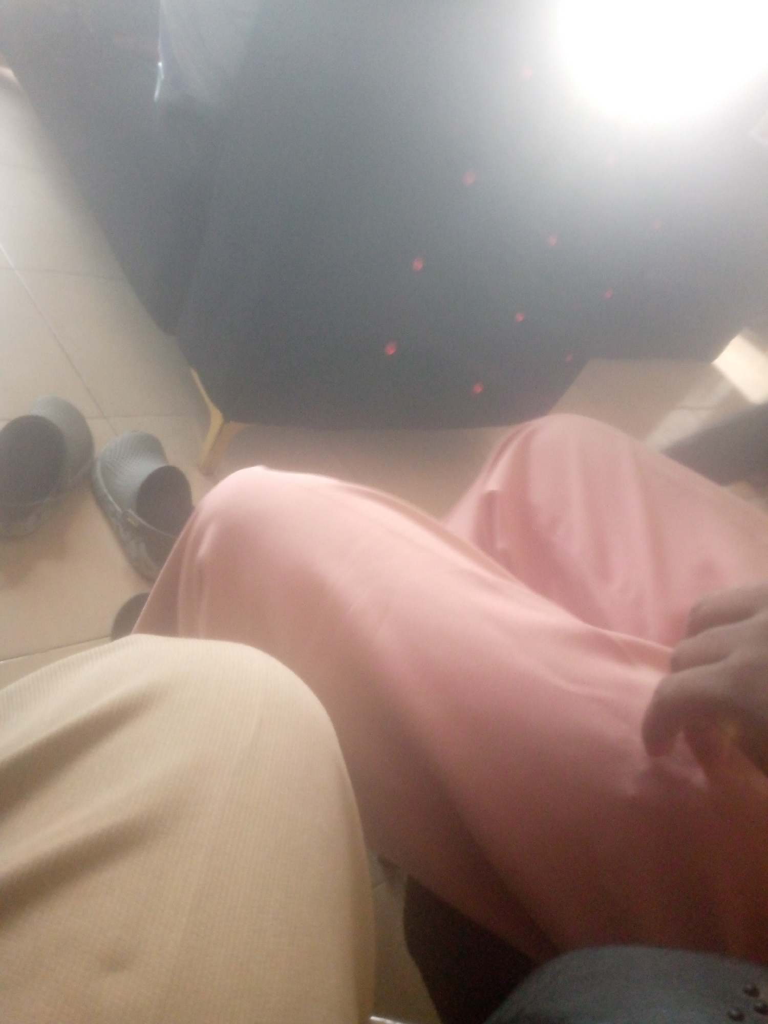 The image shows a close-up view of two individuals from the waist down, likely sitting. One person is wearing a light yellow ribbed garment, possibly a pair of pants or a skirt. The other person is draped in a flowing pink fabric, also suggesting a skirt or dress. A hand is visible resting on the pink fabric, indicating interaction. To the left, a pair of dark grey slip-on shoes or sandals are placed on a light-colored tiled floor. In the background, a dark, out-of-focus object, potentially a piece of furniture or clothing, is visible, with some red circular markings on it. The lighting is somewhat overexposed, particularly in the upper right portion of the image, suggesting an indoor setting with artificial light. The overall impression is a casual, indoor scene. Without further context or clearer details of the surroundings, it is not possible to definitively determine the location in the city of Jalingo, Nigeria. However, the tiled floor and indoor setting are common in residential and public spaces across many urban environments.